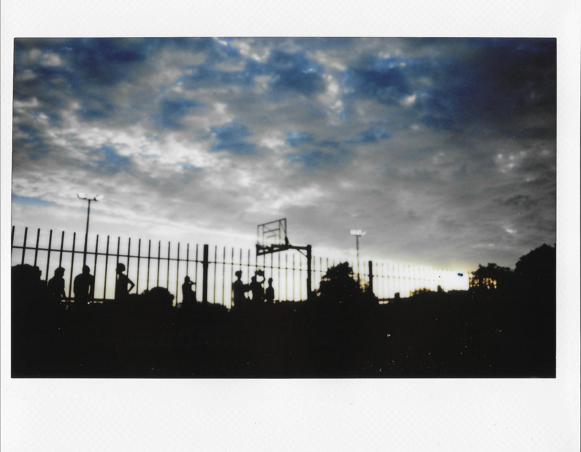 A scanned Polaroid captures silhouettes of people standing near a fence and a basketball hoop at dusk, with a dramatic cloudy sky and faint light posts in the background.