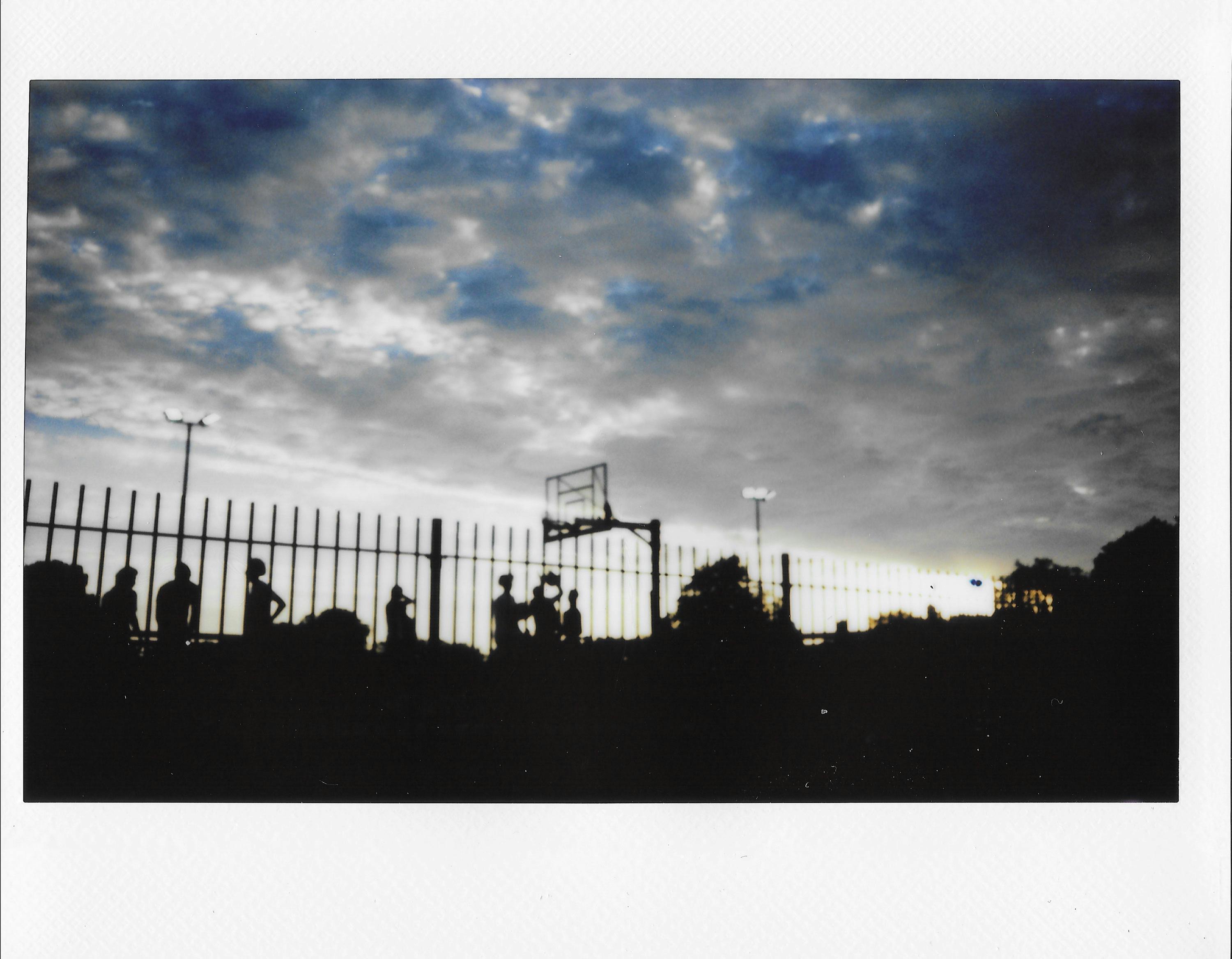 A scanned Polaroid captures silhouettes of people standing near a fence and a basketball hoop at dusk, with a dramatic cloudy sky and faint light posts in the background.