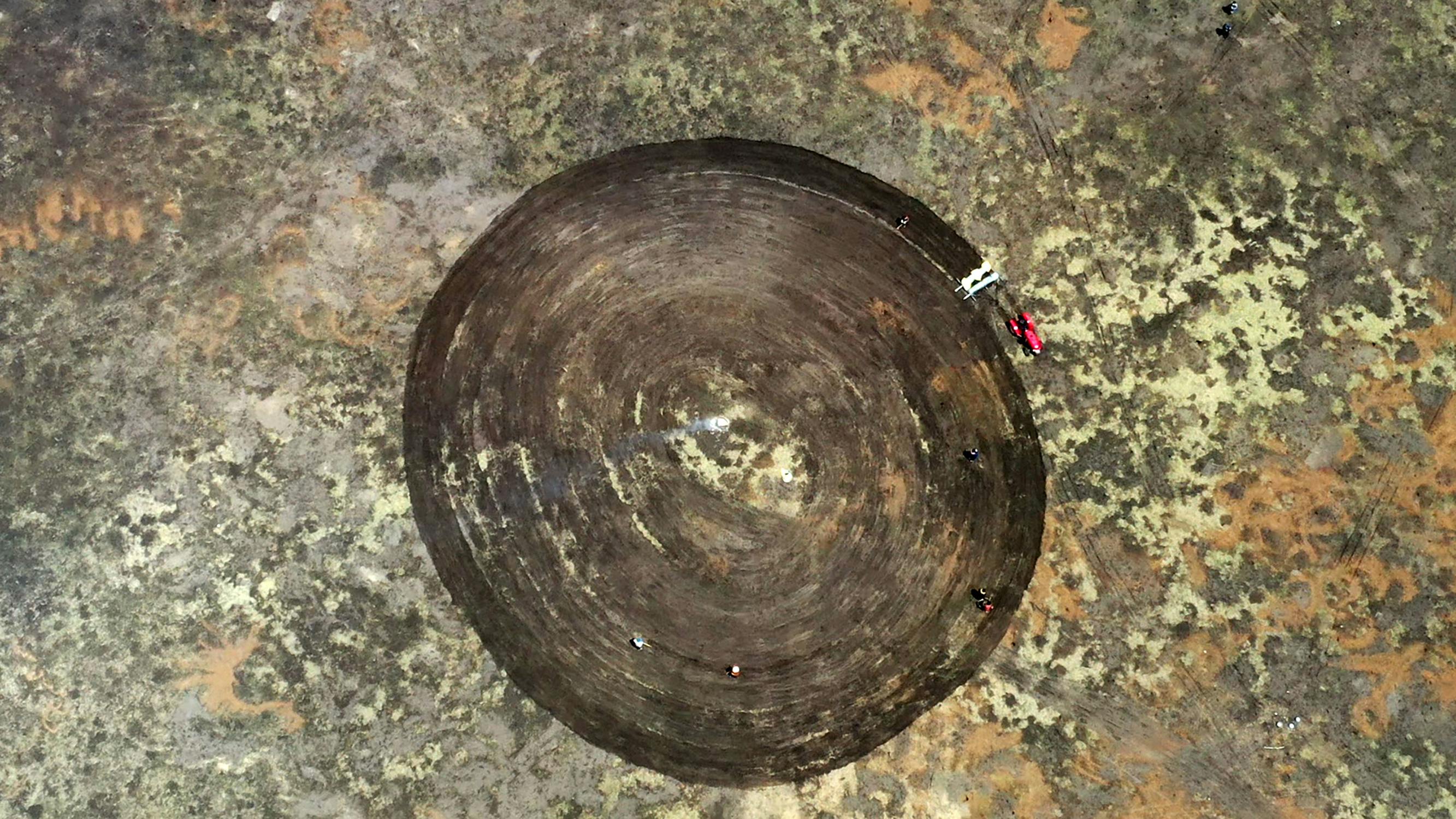 Aerial view of a large, perfectly circular patch of bare, dark soil surrounded by lighter, grassy land. Small vehicles and a few people are scattered around the edge of the circle.