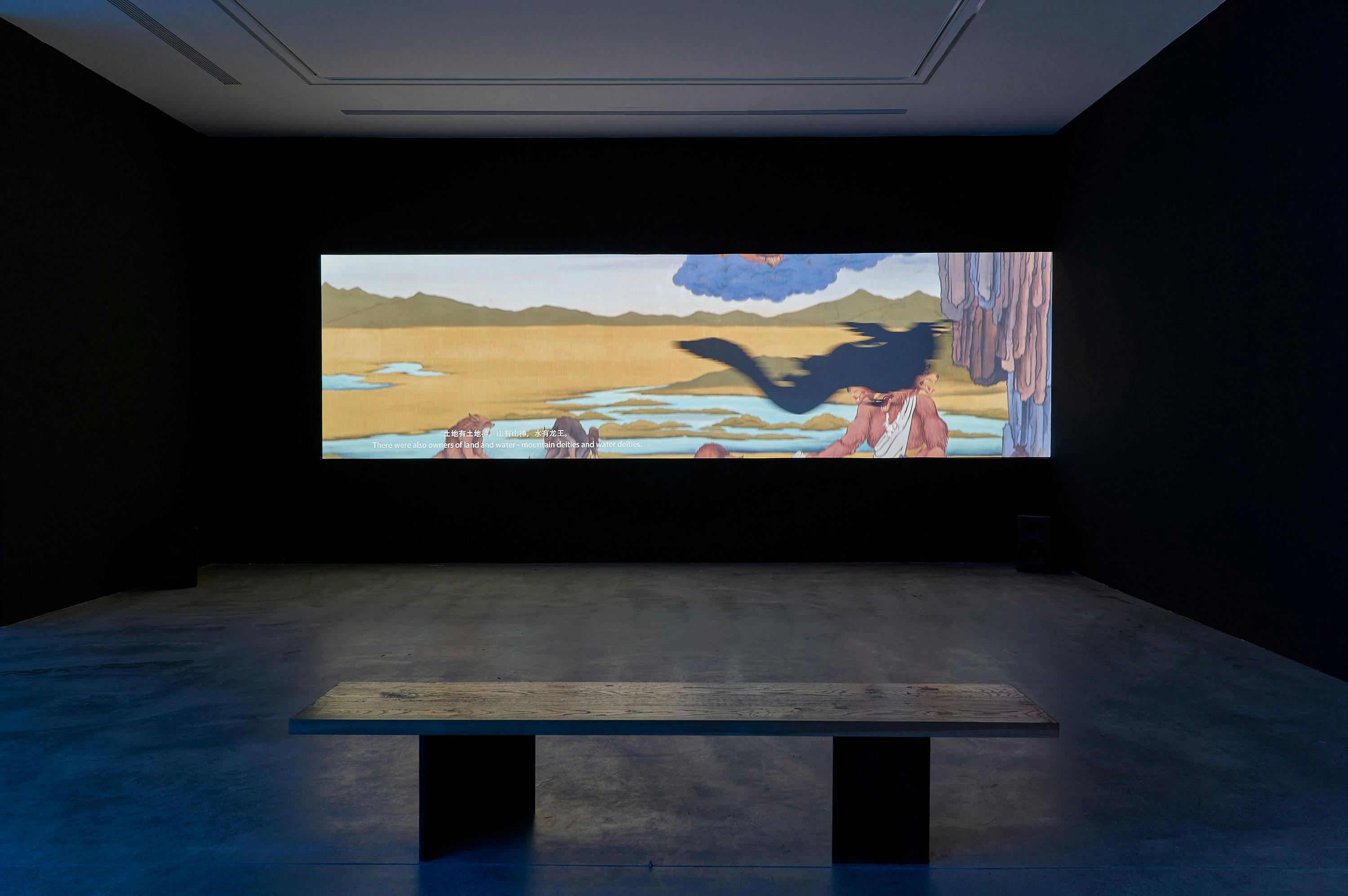A dark gallery room features a wide bench facing a large, horizontal video projection on the wall, displaying colourful surrealist art with landscapes and shadowy figures.