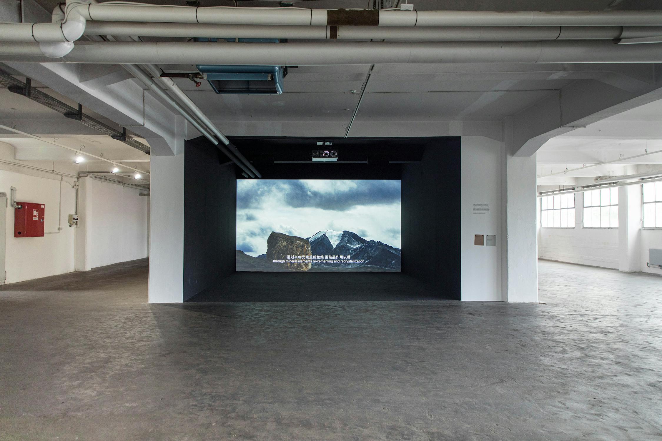 A large screen in an industrial gallery displays a video of snow-capped mountains under a cloudy sky, with subtitles in Chinese. The room has concrete floors, white walls, exposed pipes, and large windows.