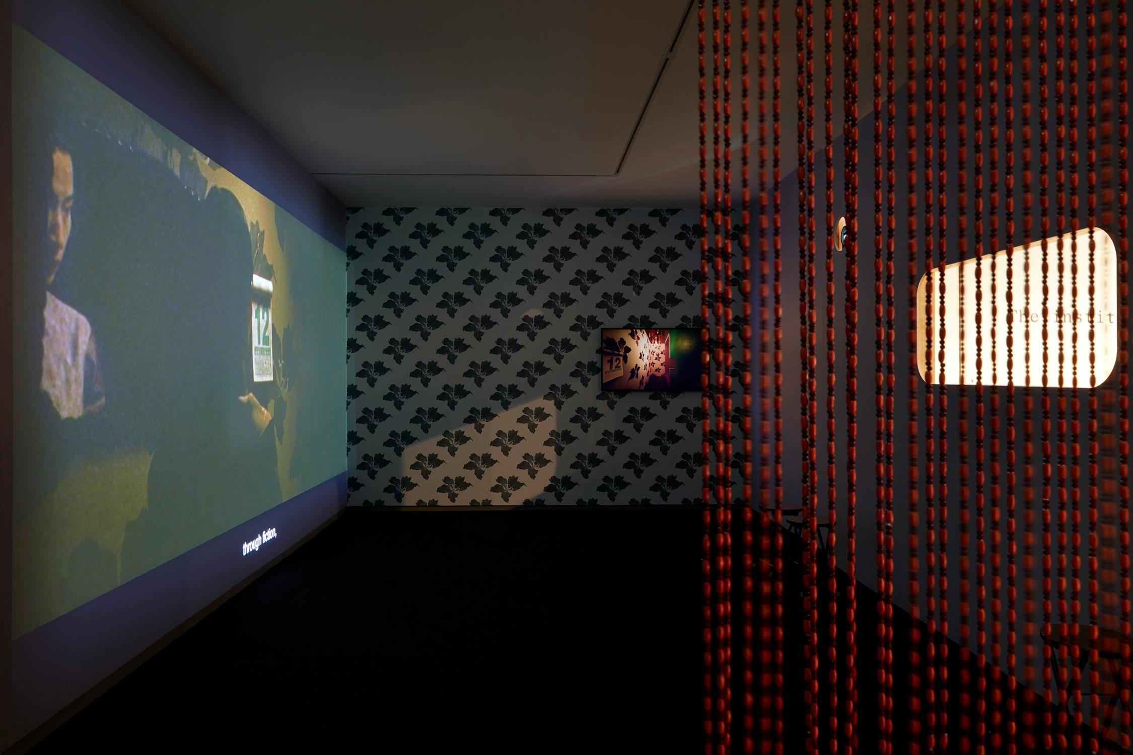 A dimly lit art gallery with a large video projection on one wall, wallpaper with a black pattern, a smaller framed artwork, and a beaded curtain partially concealing a warmly lit space.