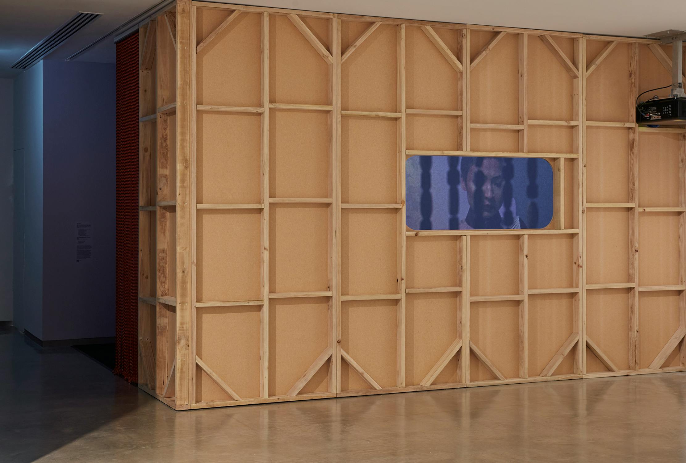 A wooden structure with exposed framing stands in a gallery. A rectangular window in the wall displays a projected video showing a person's face. The floor is polished concrete.