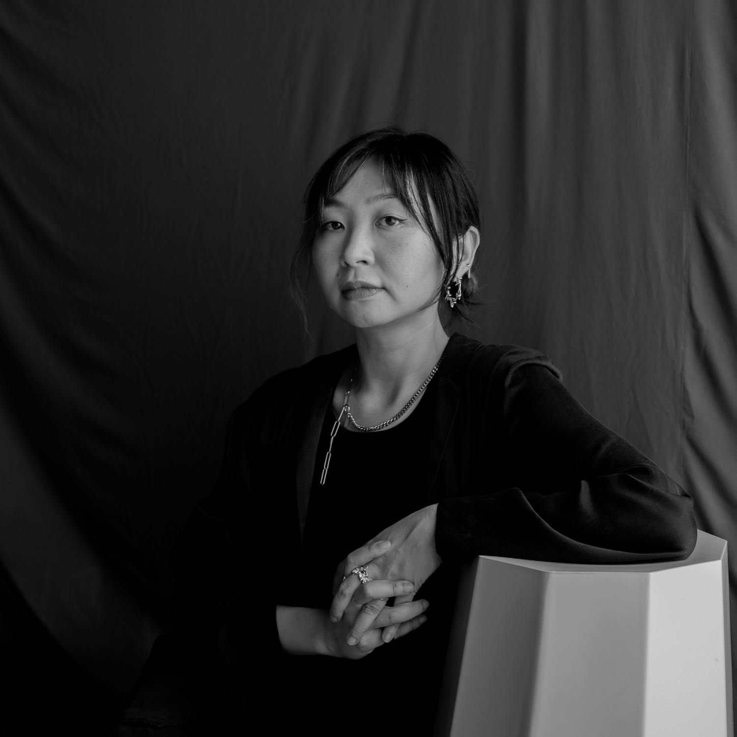 Artist Nikki Lam sits beside a chair, resting one arm on it. They wear a dark top, jewellery, and earrings. The background is a draped, dark fabric, and the image is in black and white.