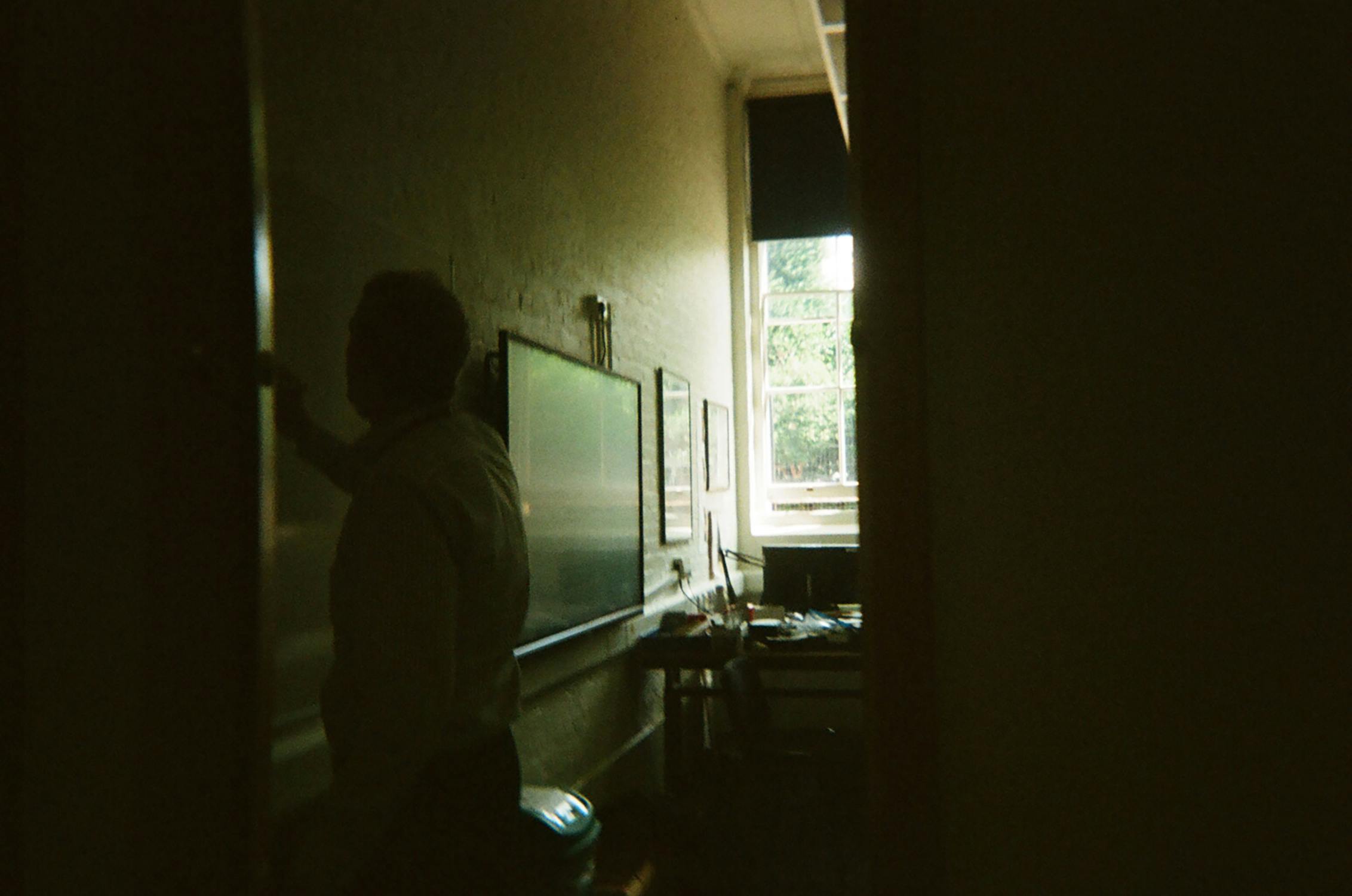 michael.'s photo shows a silhouette of a person standing near a whiteboard in a dimly lit, narrow room. Sunlight streams through a window at the far end, illuminating a desk with papers and a chair. The walls are white, and framed pictures hang beside the whiteboard.