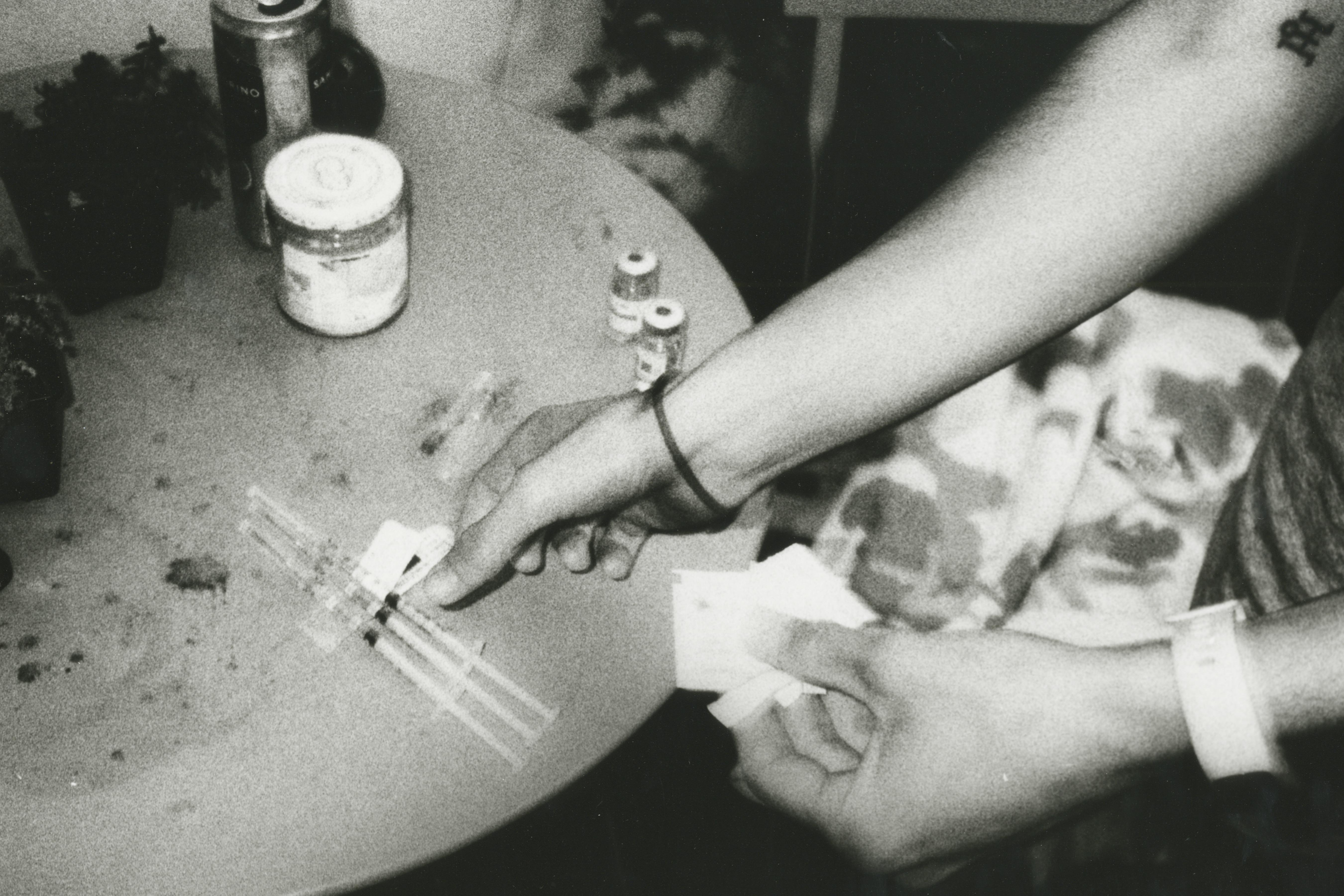 A black-and-white photo shows a person’s hands preparing testosterone shots on a round table with cards. Tins and jars are on the table. The person wears a wristband and a hair band on their wrist.