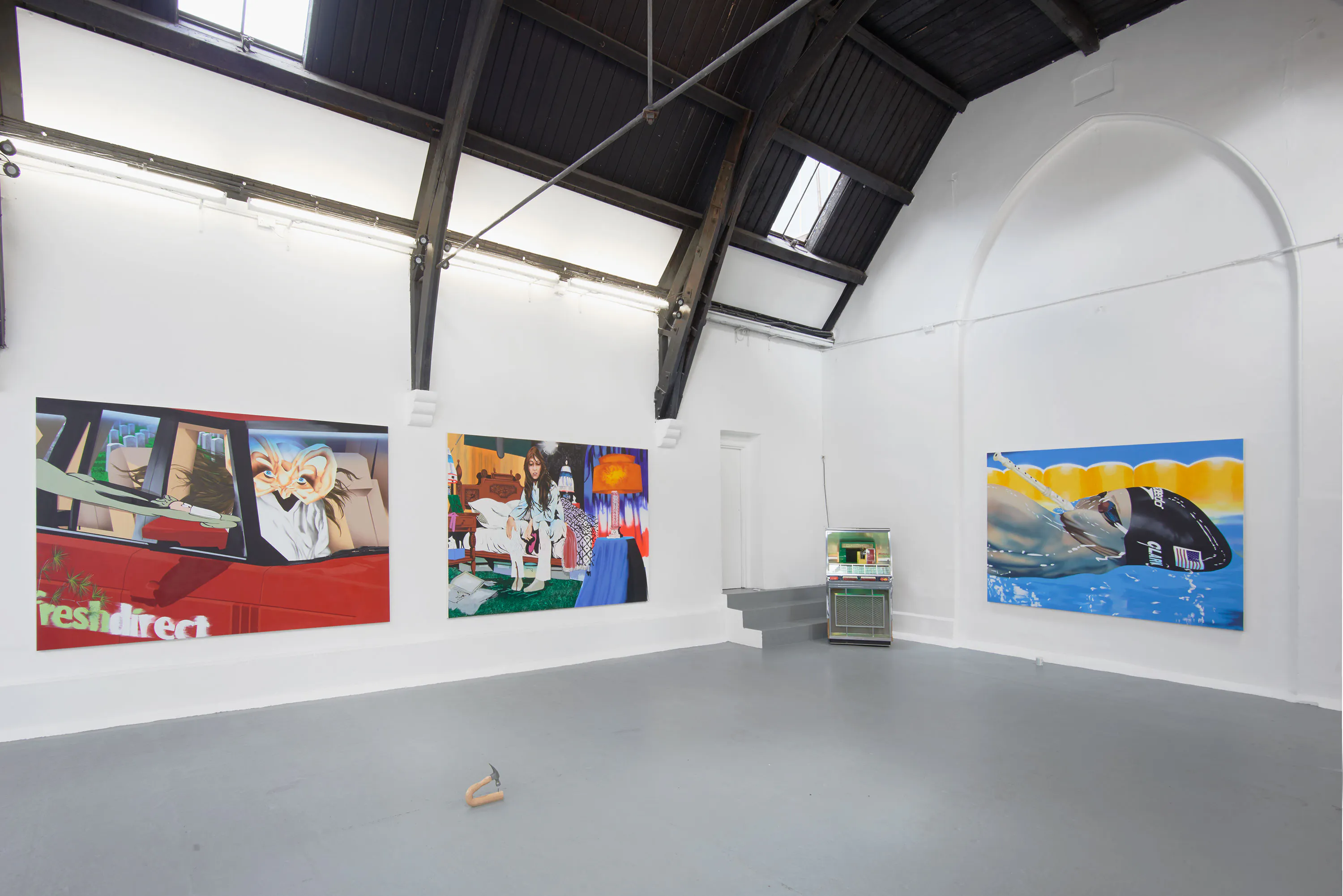A white gallery with high, dark ceilings displays three large colourful paintings by Jamian Juliano-Villani on one wall. The artworks feature abstract scenes, including a car interior, a surreal dining table, and a swimmer in blue water. A small fridge sits beside the paintings on a grey floor.