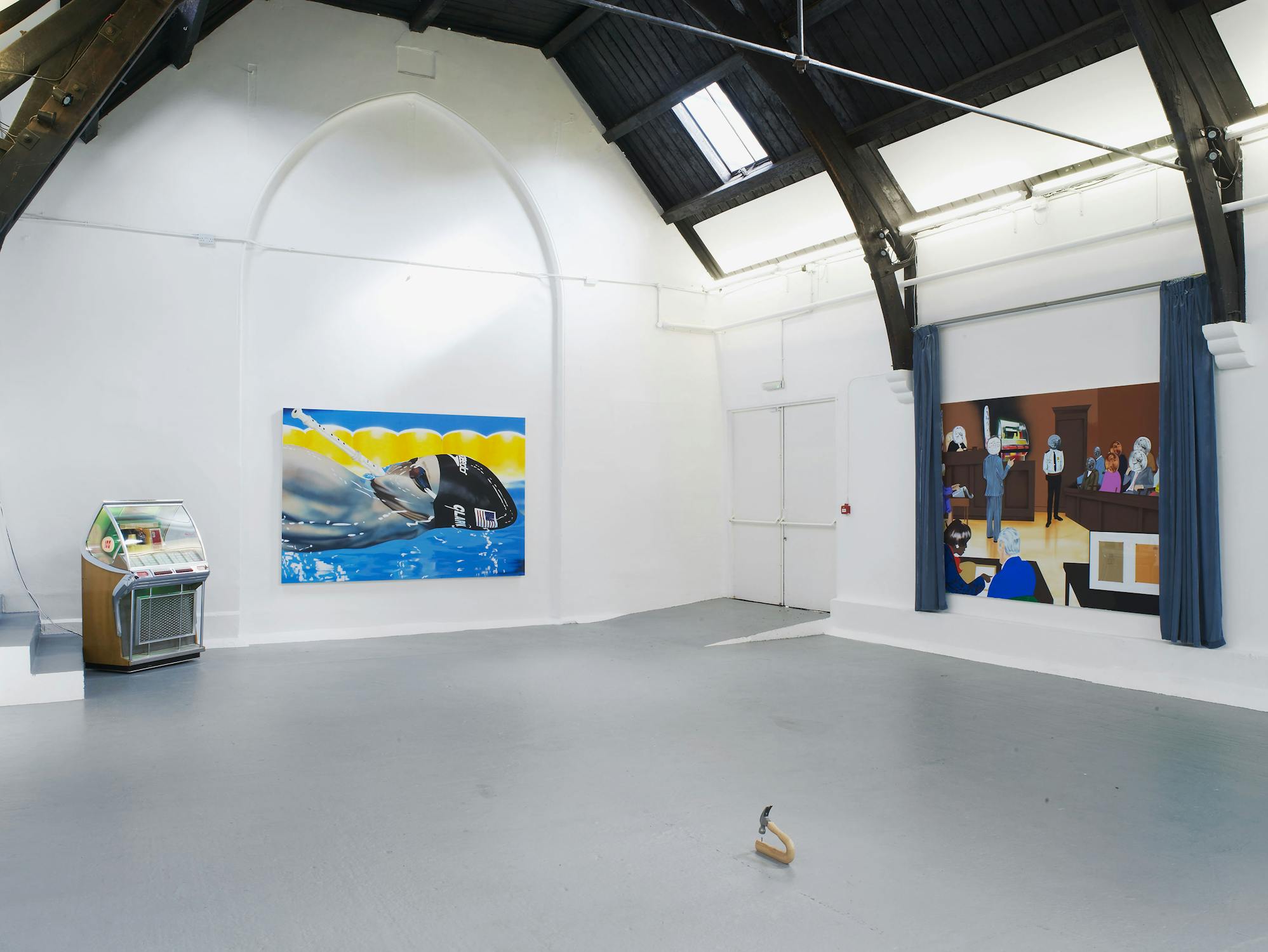 A bright art gallery with high ceilings and skylights displays a colourful painting of a swimmer submerged in water on one wall and a large artwork of people in a room on another. A jukebox stands to the left, and a small wooden object sits on the grey floor in the centre.