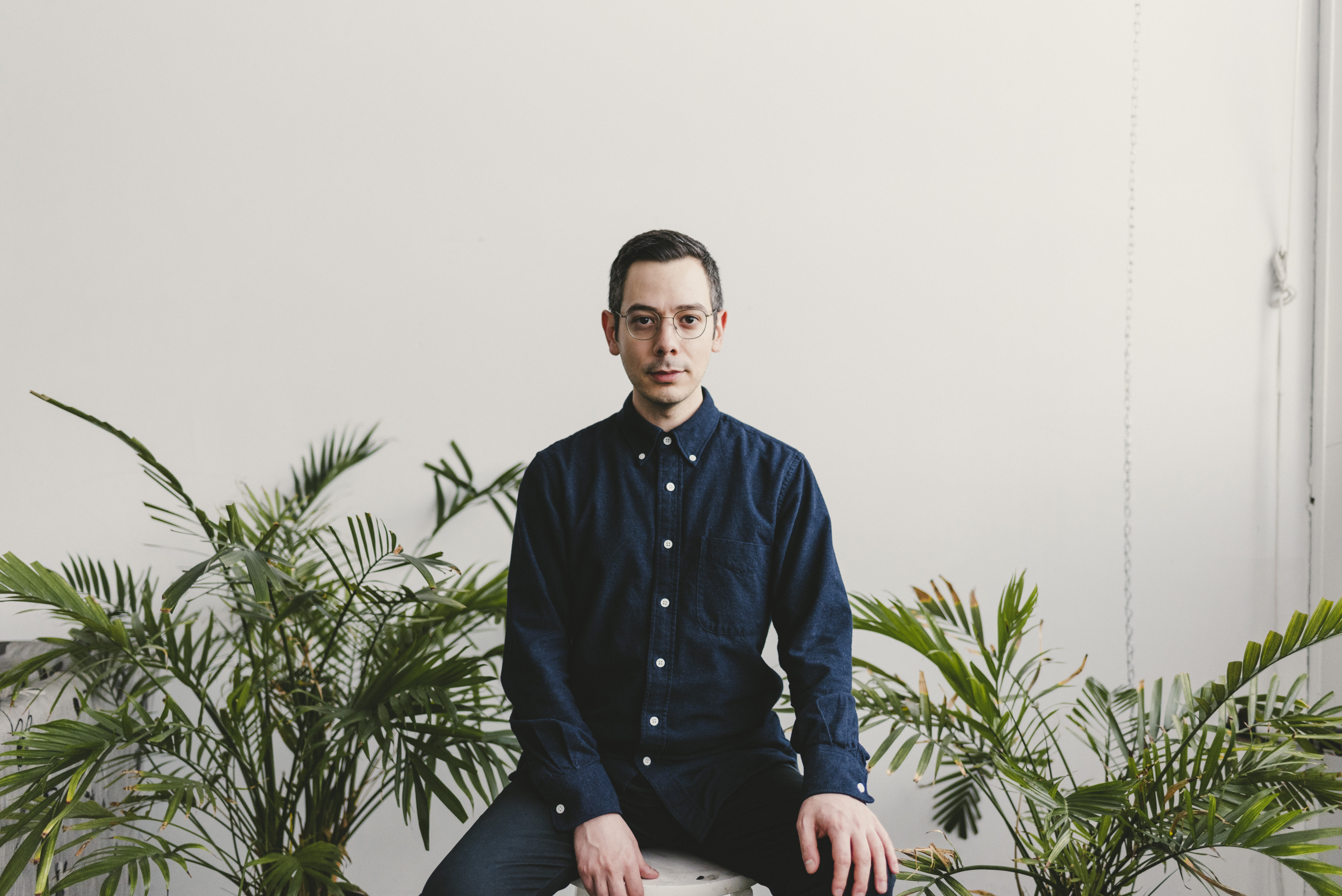 Portrait of Erdem Taşdelen. A man with short dark hair and glasses sits on a stool, facing forward, wearing a dark blue button-up shirt and jeans. He is surrounded by green potted plants against a plain white wall in a bright, minimalistic room.
