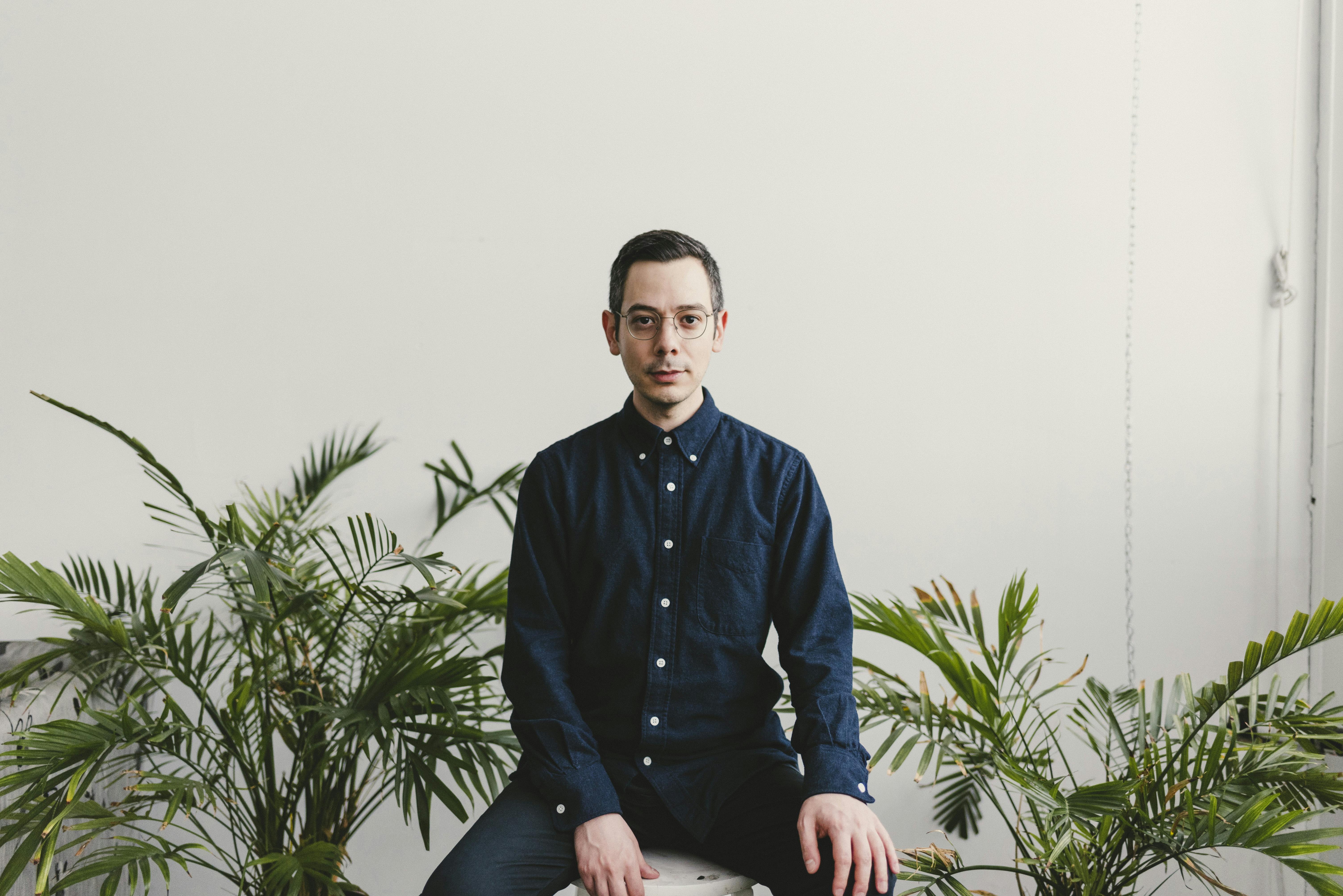 Portrait of Erdem Taşdelen. A man with short dark hair and glasses sits on a stool, facing forward, wearing a dark blue button-up shirt and jeans. He is surrounded by green potted plants against a plain white wall in a bright, minimalistic room.