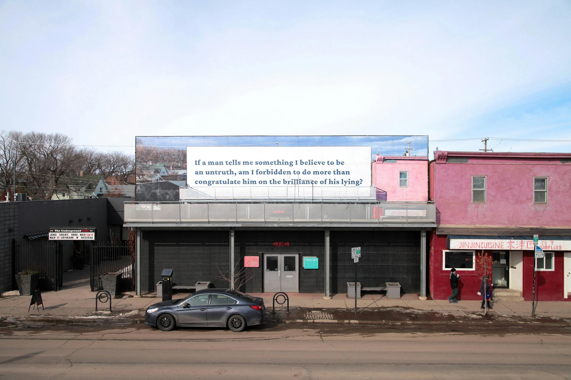A grey car is parked on a street in front of a closed building with a large billboard above. The billboard reads, “If a man tells me something I believe to be an untruth, am I forbidden to do more than congratulate him on the brilliance of his lying?” Two people walk nearby.