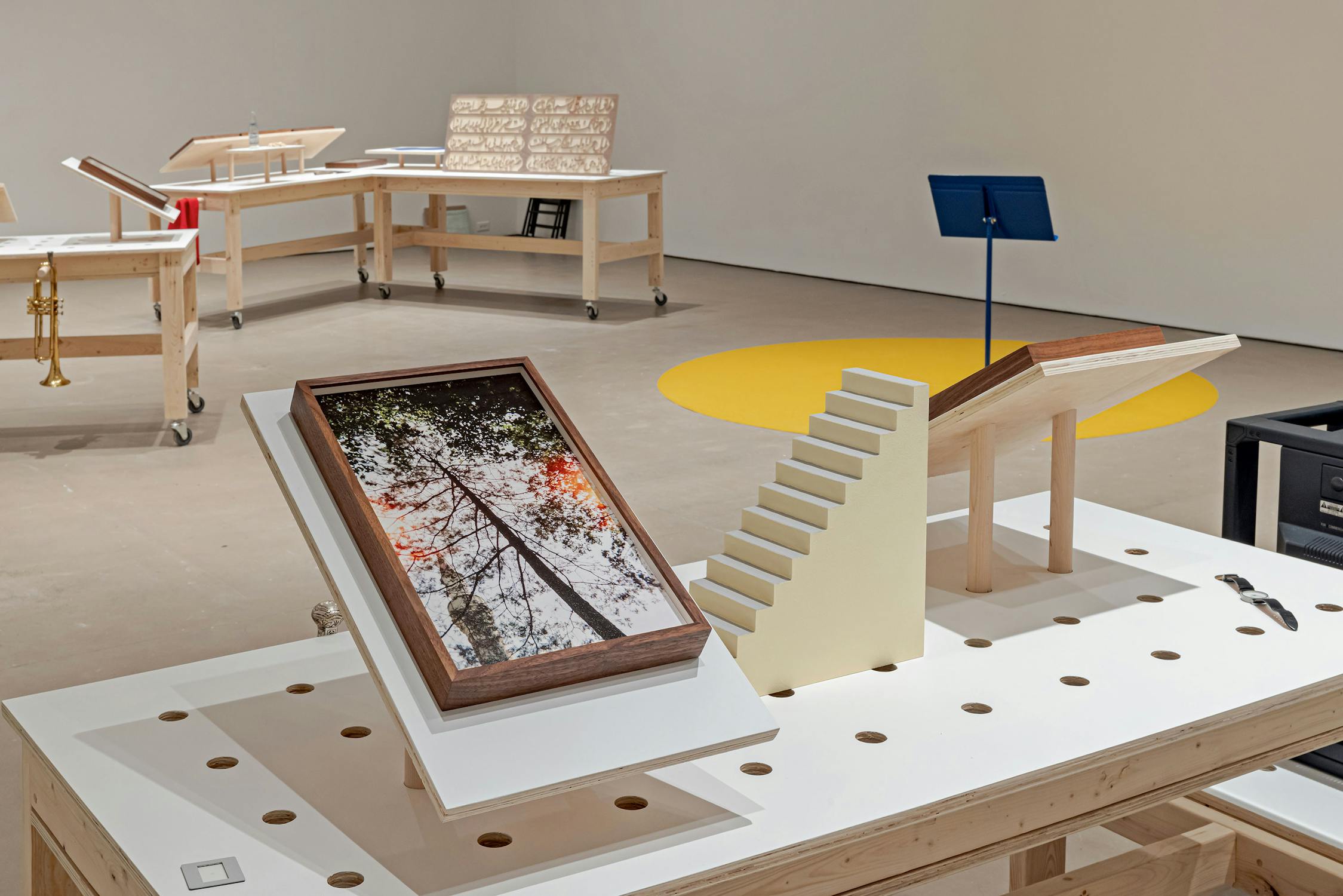 A modern art exhibition features angled wooden tables with objects: a framed photograph of tall trees, a small model staircase, and music stands. In the background are more tables with books and a circular yellow carpet on the floor. The space is minimal and well-lit.