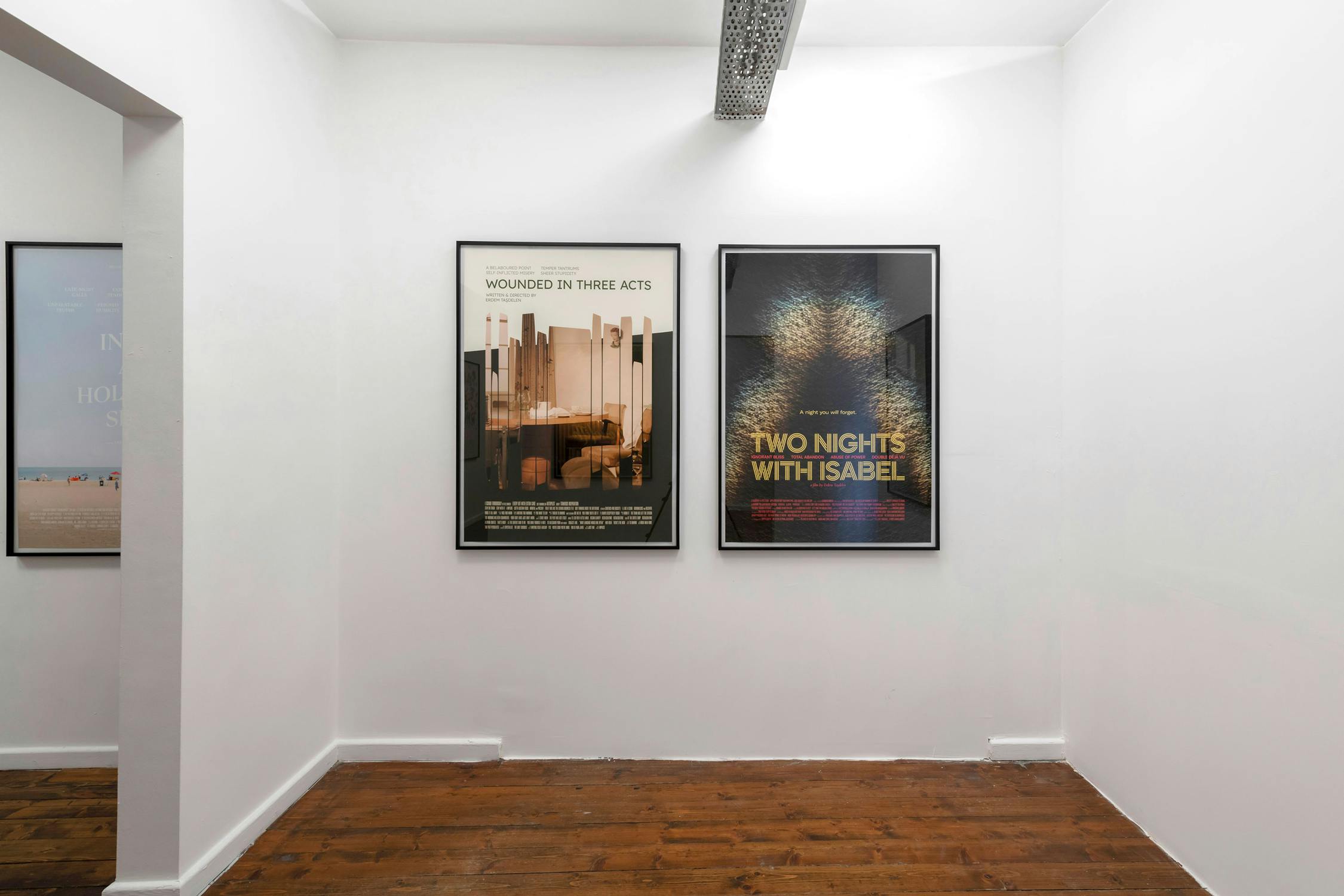A small white-walled room with wooden floors displays two framed posters on the wall. The left poster shows abstract beige structures, whilst the right poster features colourful, blurry lights and text reading “TWO NIGHTS WITH ISABEL.”.