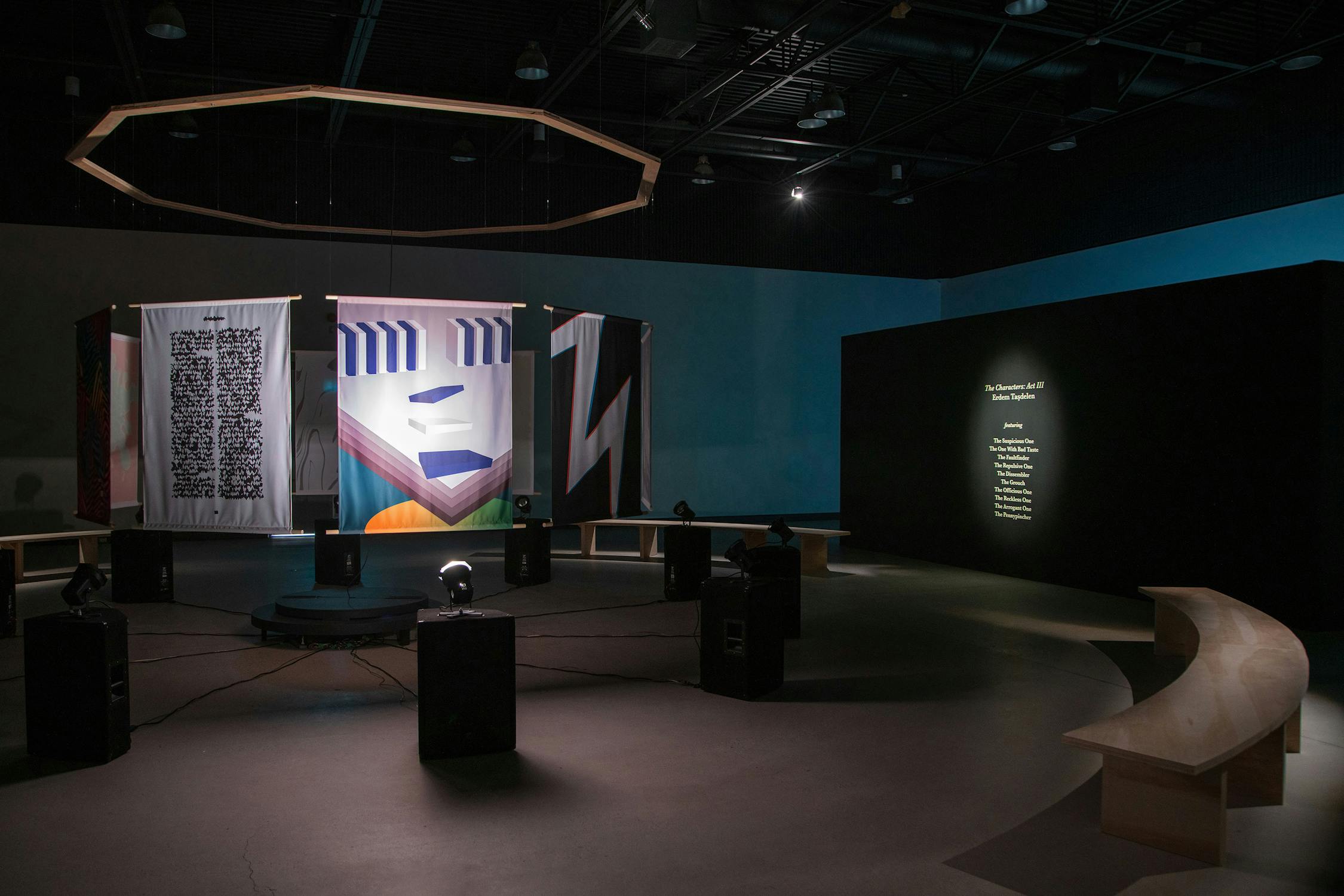 A dimly lit gallery with abstract banners, geometric designs, and a circular platform. Several spotlights and speakers face the platform. A curved bench is in the foreground, and a glowing text panel is on the dark wall to the right. The ceiling is high and industrial.