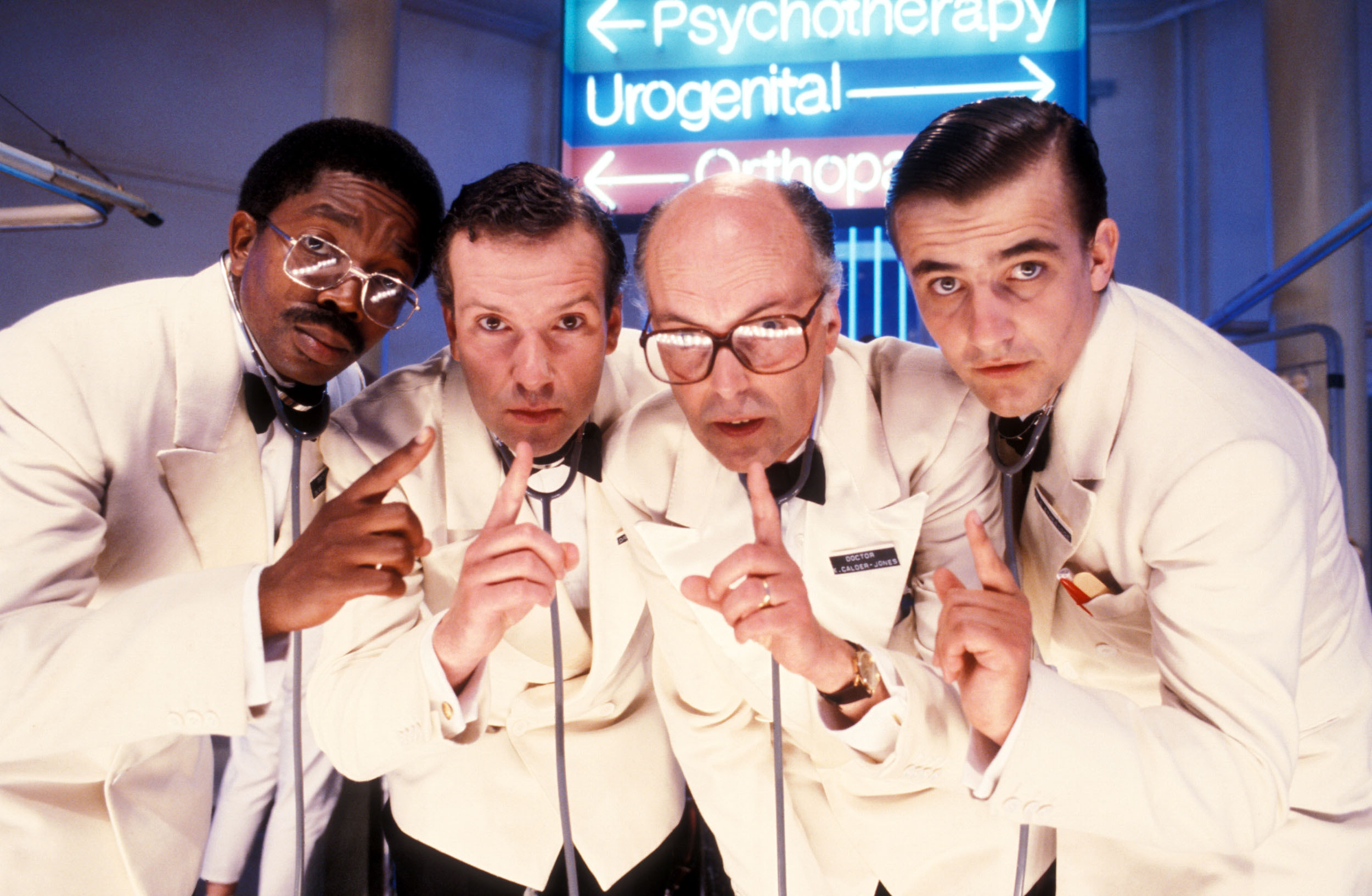 Four men in white lab coats lean forward, holding up index fingers as if to signal quiet. They wear stethoscopes and serious expressions. Behind them, blue and pink neon hospital signs read “Psychotherapy,” “Urogenital,” and “Orthopaedics”.
