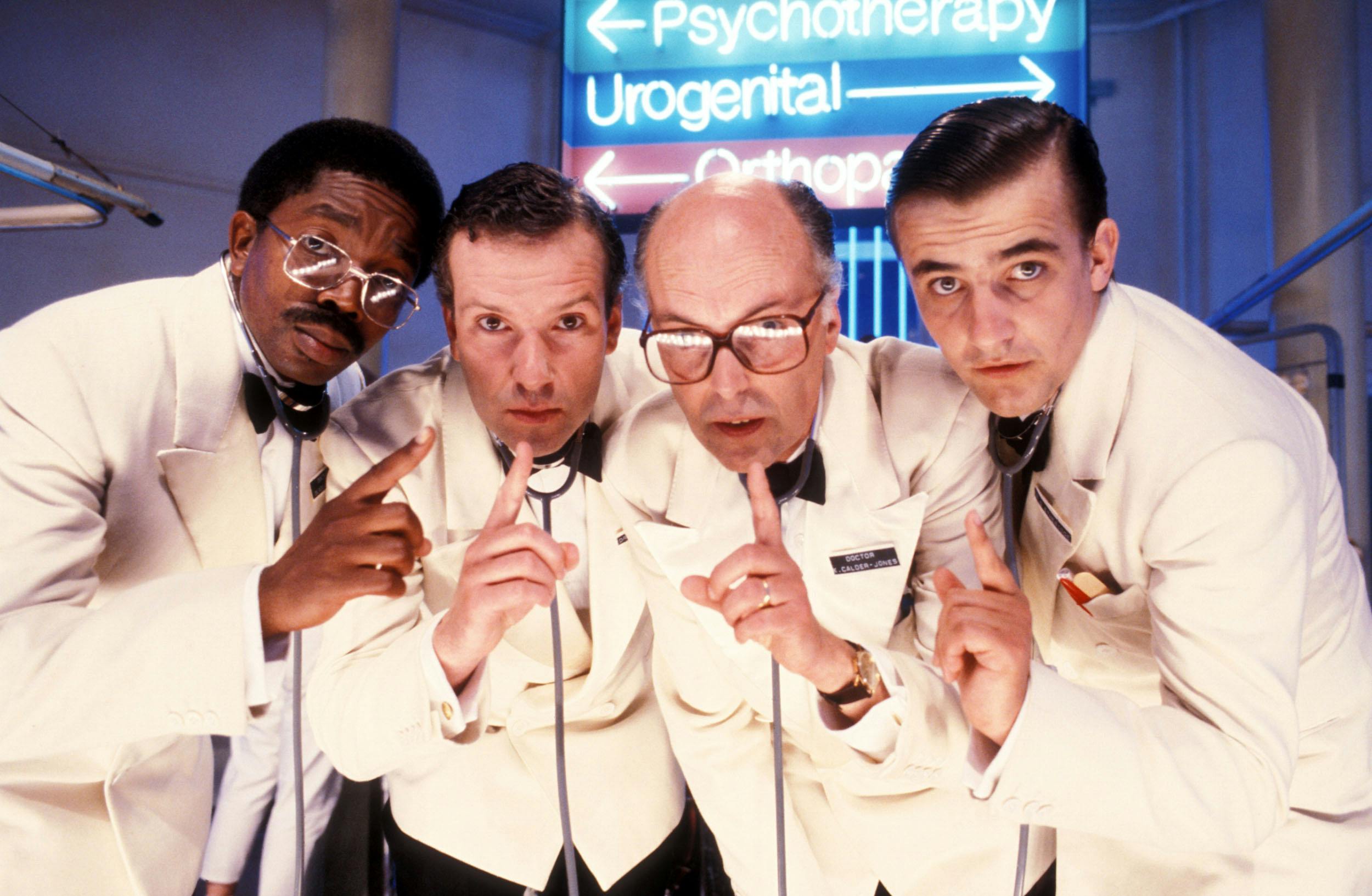 Four men in white lab coats lean forward, holding up index fingers as if to signal quiet. They wear stethoscopes and serious expressions. Behind them, blue and pink neon hospital signs read “Psychotherapy,” “Urogenital,” and “Orthopaedics”.