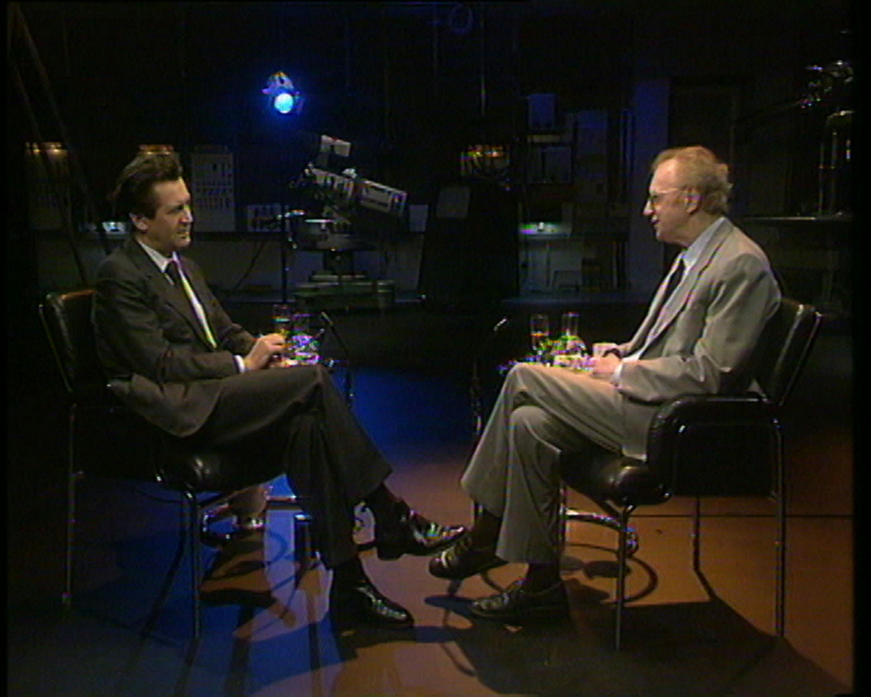 Melvyn Bragg and Dennis Potter facing each other in black chairs on a dimly lit TV studio set. They have drinks on a small table between them. A large camera and blue spotlight are visible in the background, creating a serious interview atmosphere.