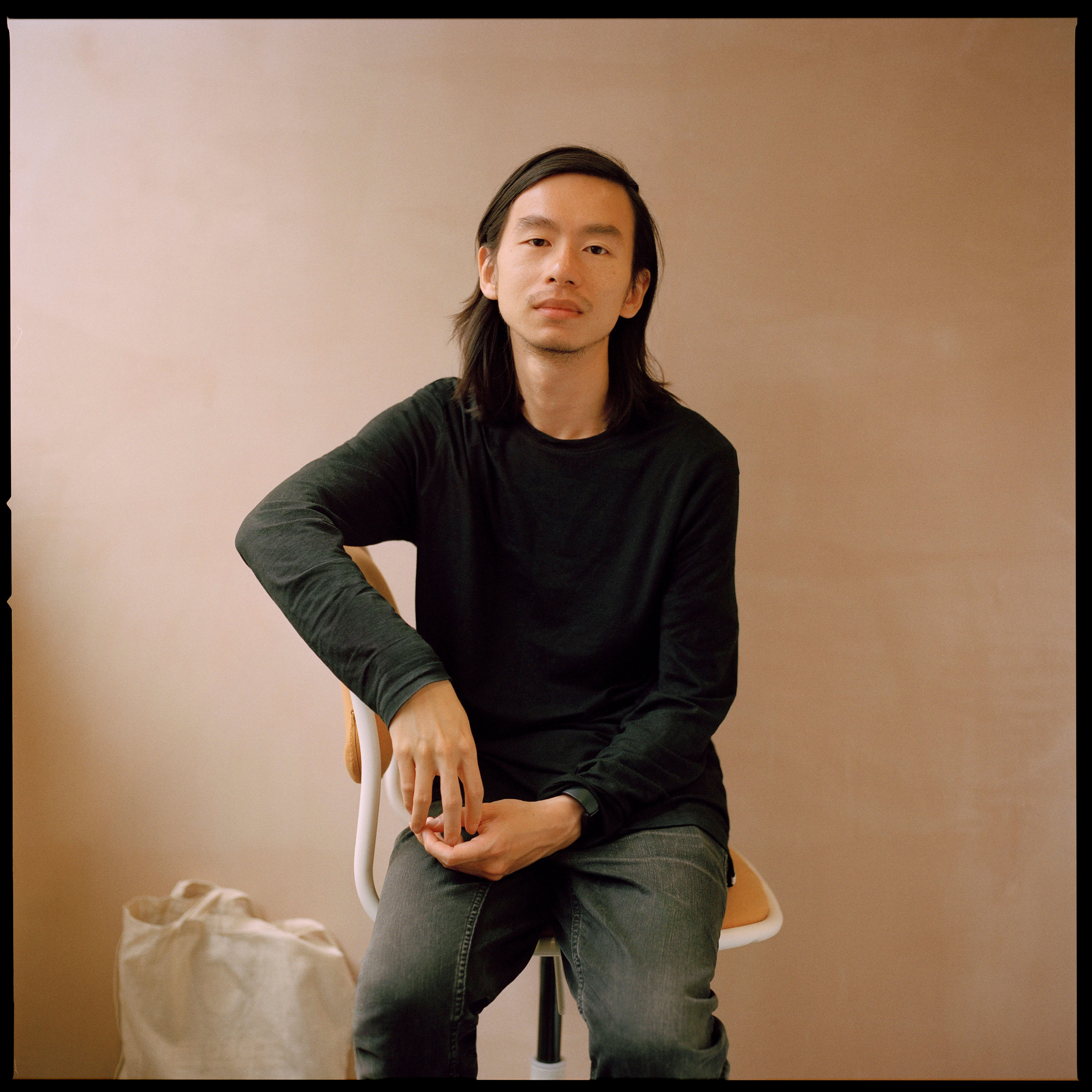 A portrait of Chris Zhongtian Yuan, sitting on a chair against a plain beige wall, wearing a black long-sleeved shirt and grey jeans. One hand rests on their knee while the other hand drapes over the chair's back. A white bag sits on the floor nearby.