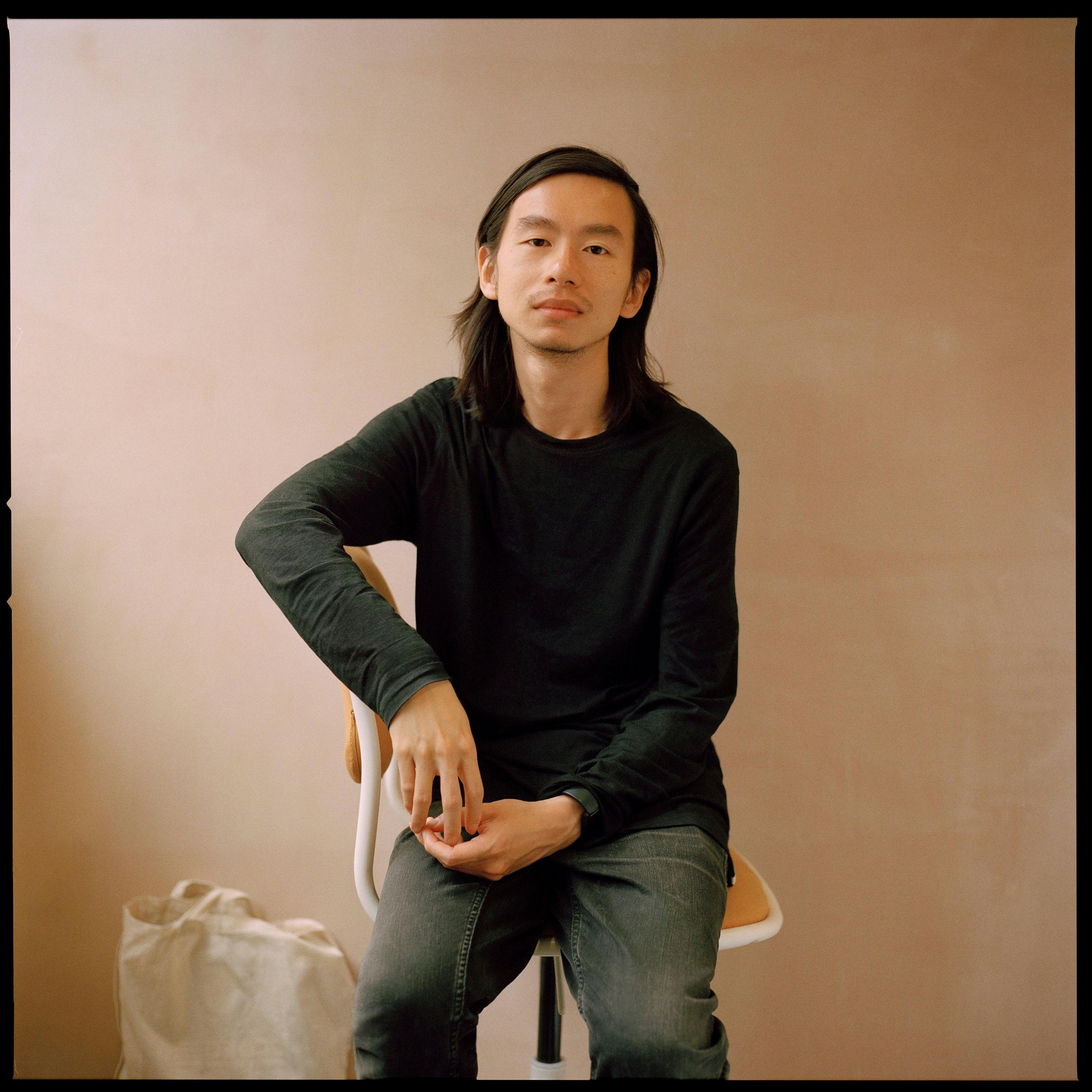 A portrait of Chris Zhongtian Yuan, sitting on a chair against a plain beige wall, wearing a black long-sleeved shirt and grey jeans. One hand rests on their knee while the other hand drapes over the chair's back. A white bag sits on the floor nearby.