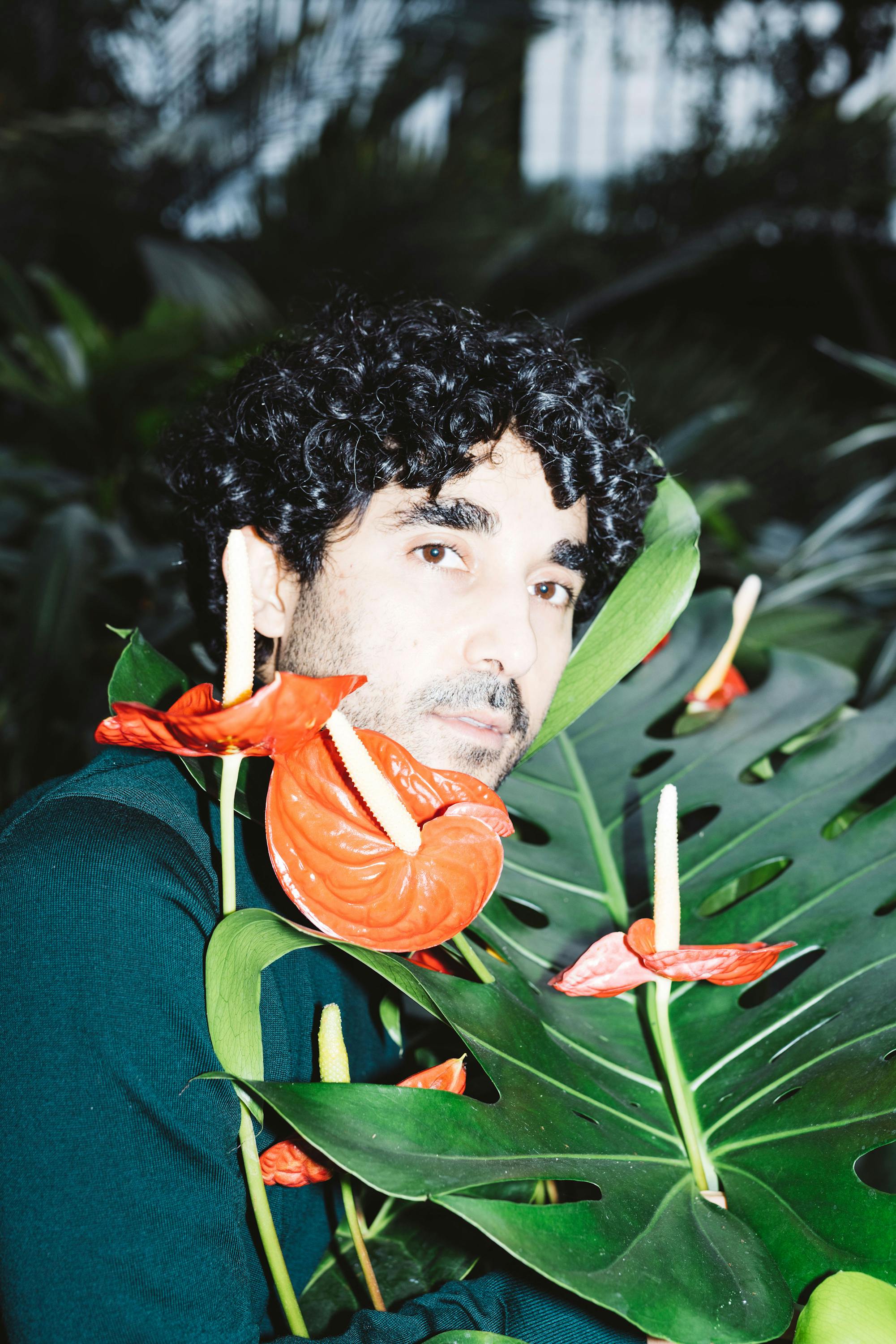 A portrait of Murat Adash with curly dark hair and a short beard gazes forward, surrounded by bright red anthurium flowers and large green monstera leaves. The background has a lush, tropical feel with blurred greenery. The lighting is bright, giving a surreal vibrant effect.