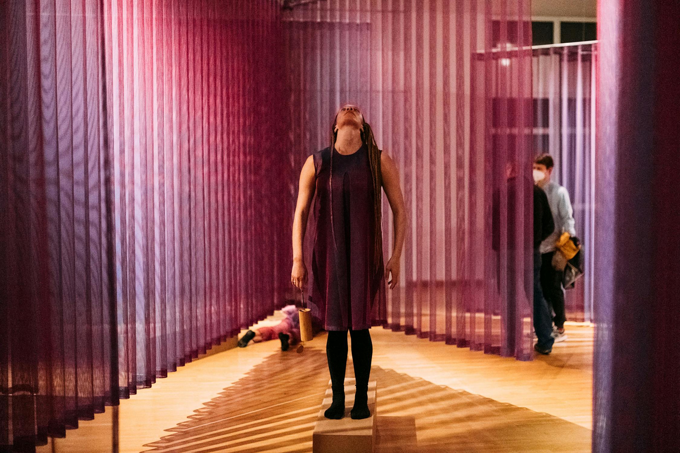 A woman in a sleeveless purple dress stands on a small platform, head tilted back, in a room with sheer purple curtains. A child lies on the floor in the background, and a person wearing a mask walks by on the right. The setting has warm lighting and a wooden floor.