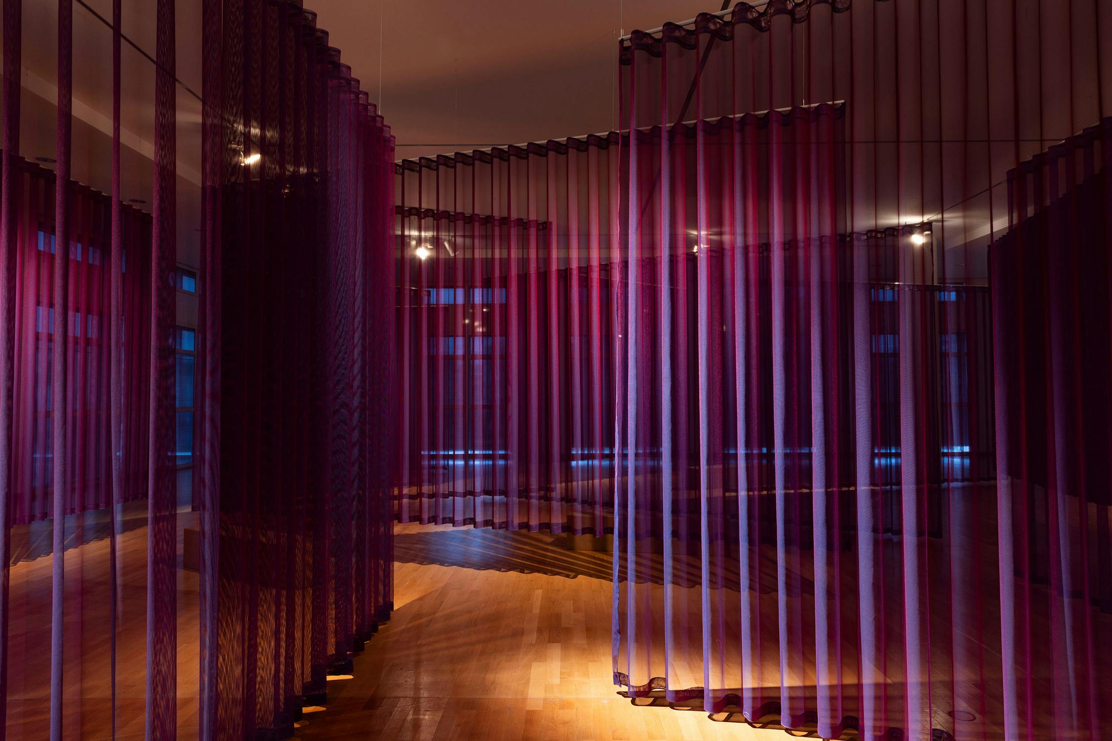 A room with wooden floors is filled with tall, semi-transparent purple and burgundy fabric curtains hanging in curved, wavy lines. Spotlights from the ceiling cast soft light, creating overlapping shadows and a layered, abstract, immersive environment.