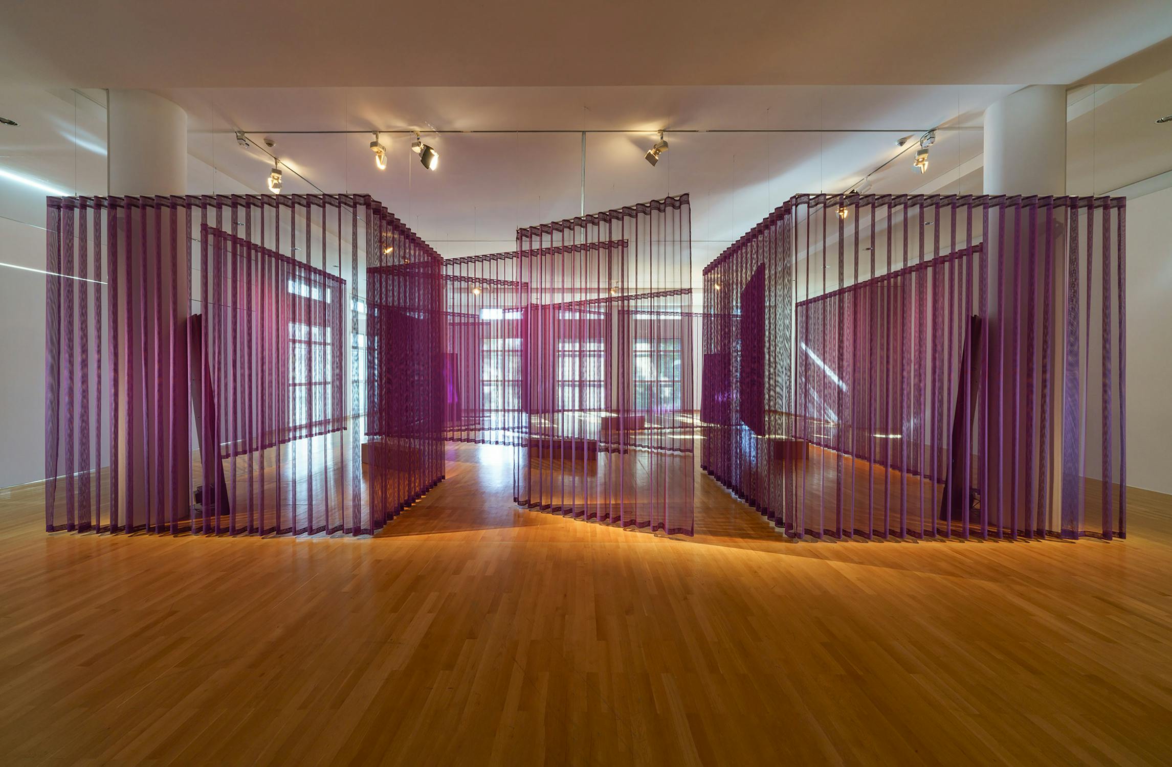 A modern art installation by Murat Adash in a gallery features tall, translucent purple panels arranged in zigzag patterns on a polished wooden floor. Bright ceiling lights cast soft shadows, while a mirrored back wall reflects the geometric structure and windows beyond.