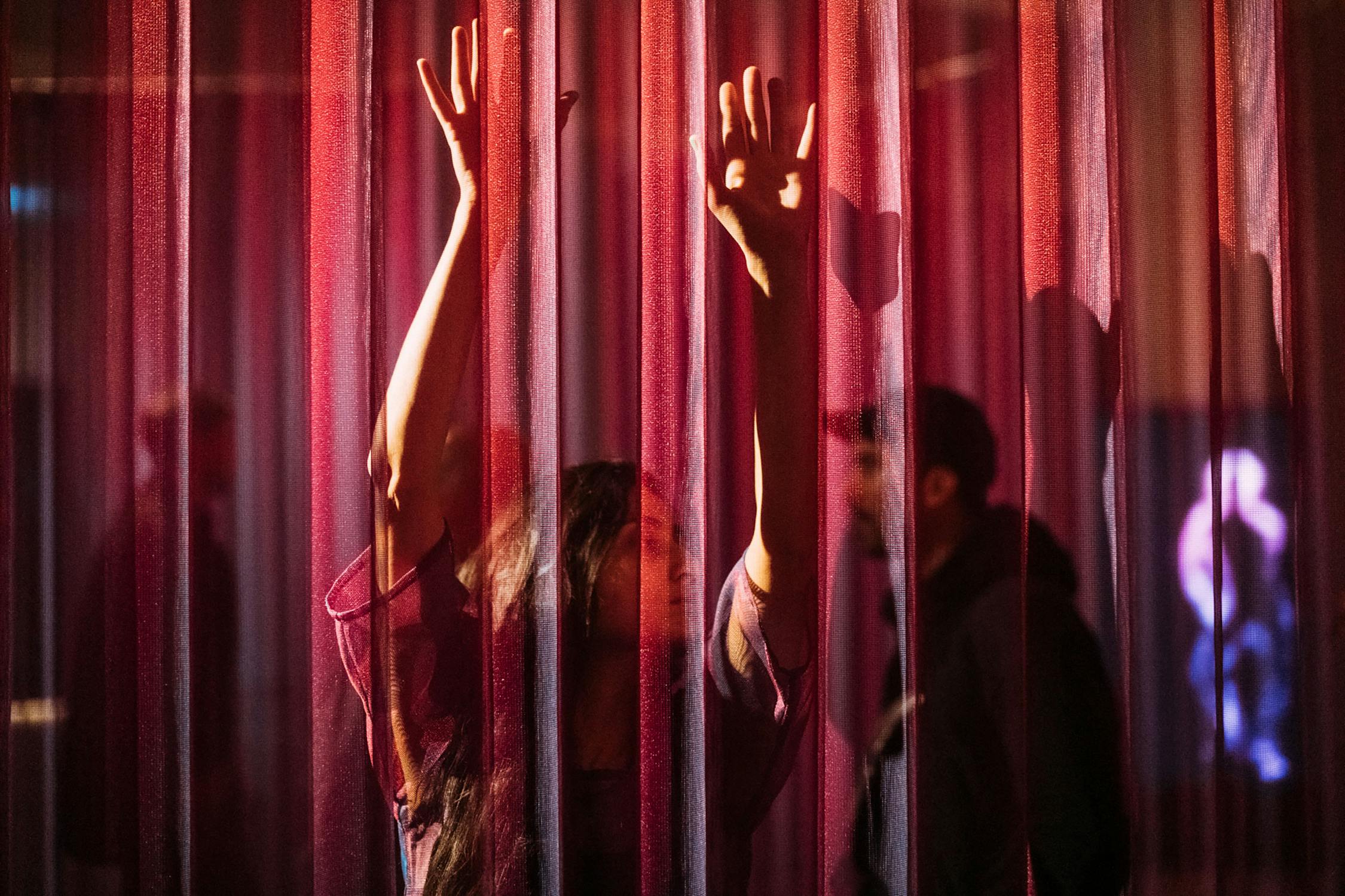 A woman with raised arms stands behind a sheer, red-lit curtain, her face and hands pressed towards the fabric. In the background, a silhouette of a person is visible through the curtain, creating an atmospheric, dramatic scene with warm lighting.