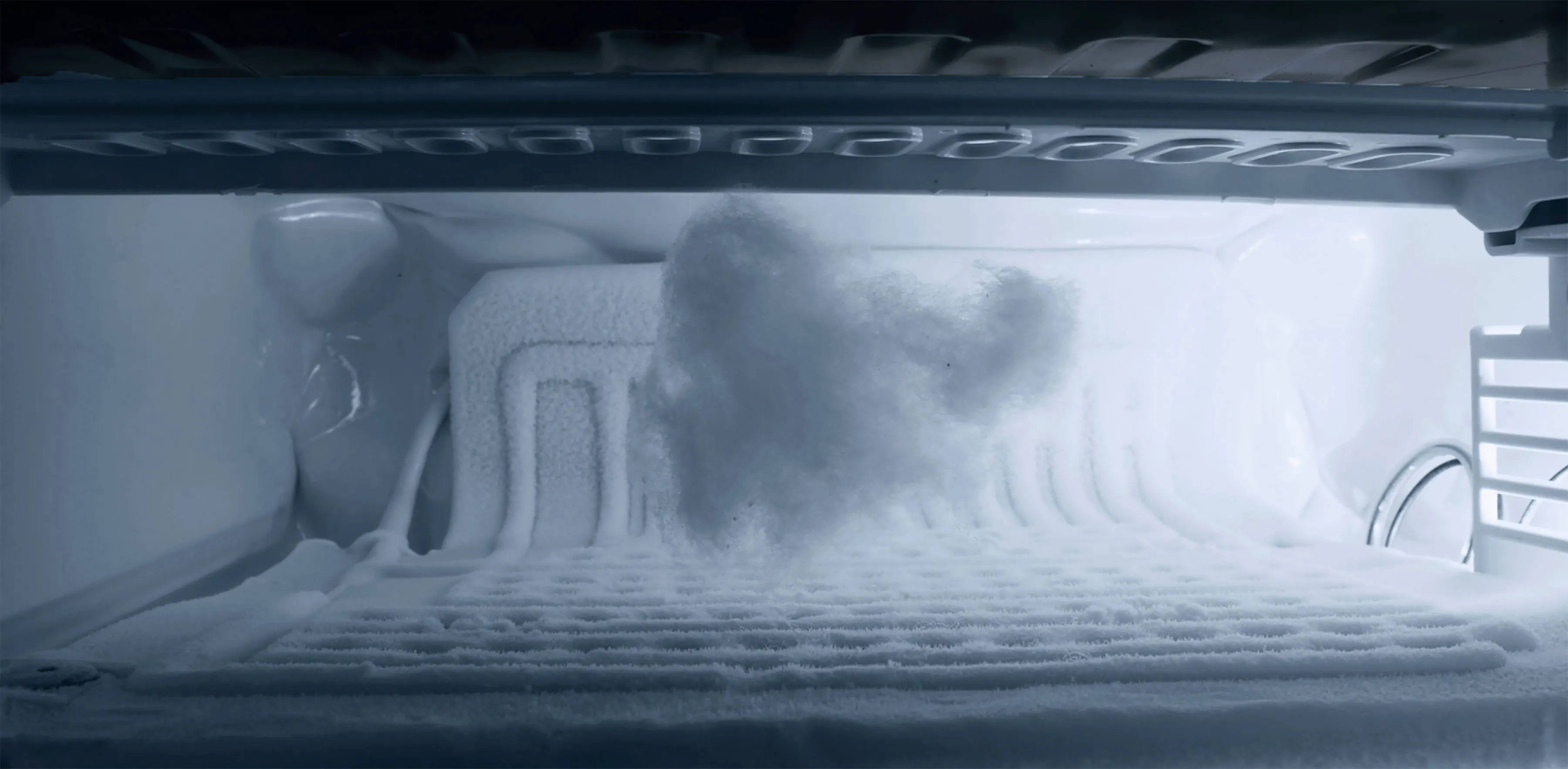Close-up view inside a freezer with a thick build-up of white frost and ice along the back and bottom surfaces. A large, fluffy clump of frost is in the centre, illuminated by a cold blue-tinted light. The freezer appears empty otherwise.