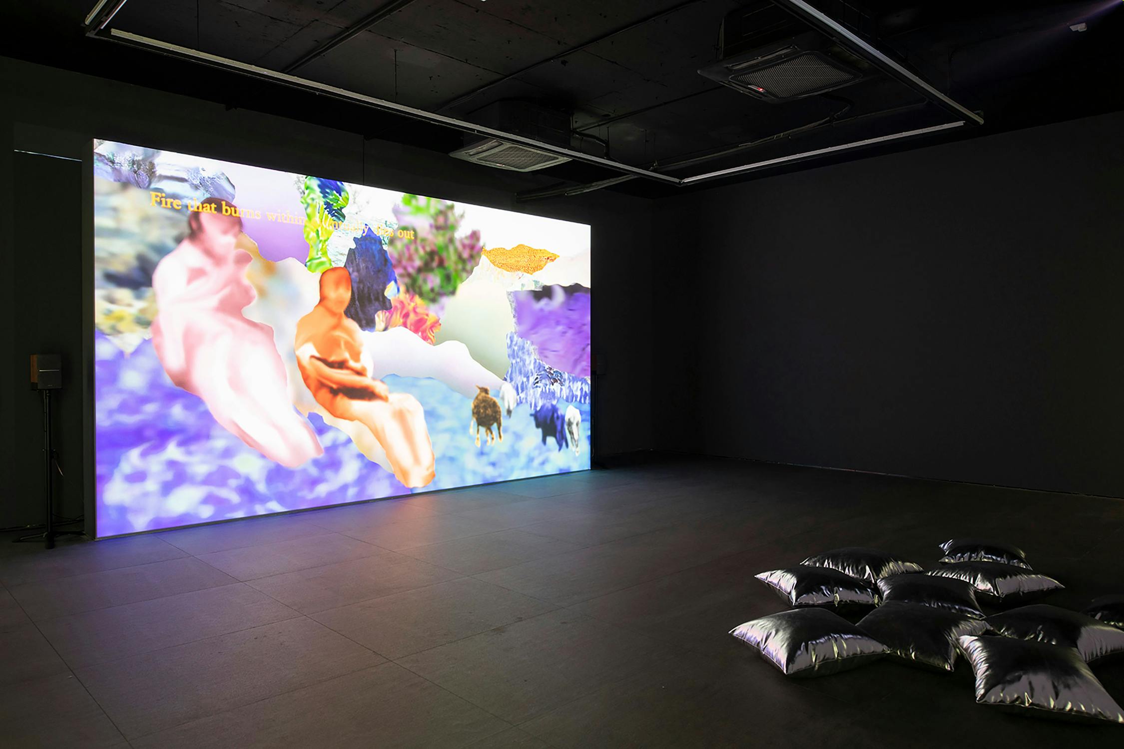 A dark gallery room features a brightly lit digital projection of two abstract, human-like figures in pastel colours among dreamy, surreal landscapes. Nearby, a cluster of silver metallic cushions is arranged on the floor for seating or lounging.