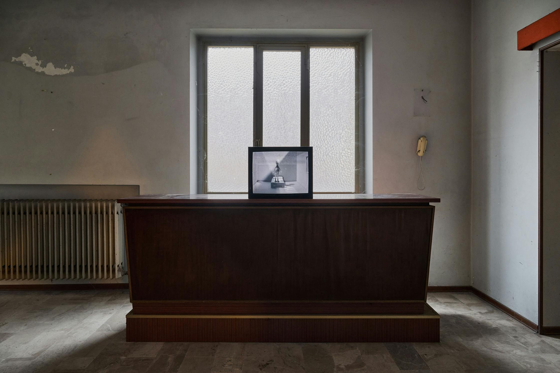 A dimly lit, empty room features a wooden reception desk facing a window with frosted glass. On the desk sits a framed black and white photo. To the right, a yellow wall telephone is mounted, and a radiator is visible on the left wall. The walls appear worn.