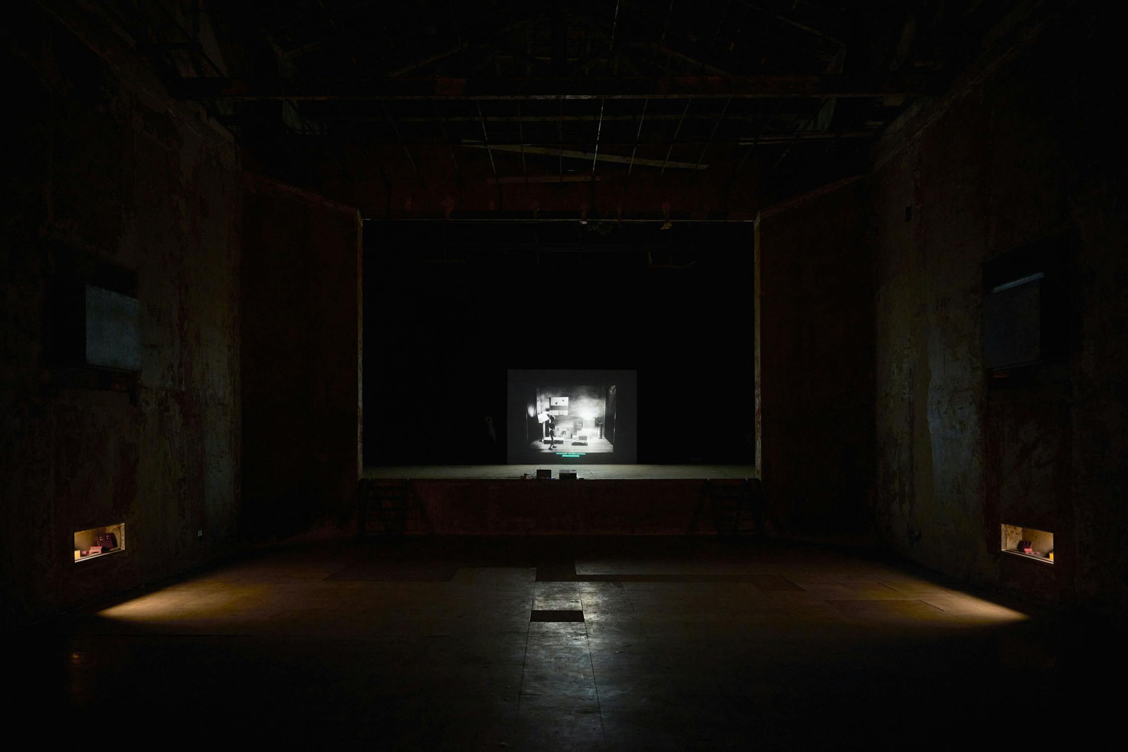 A dark, empty theatre with rough, unfinished walls and minimal lighting. At the centre stage, a black-and-white image or film is projected, showing a dim room with furniture. The atmosphere is eerie and abandoned.