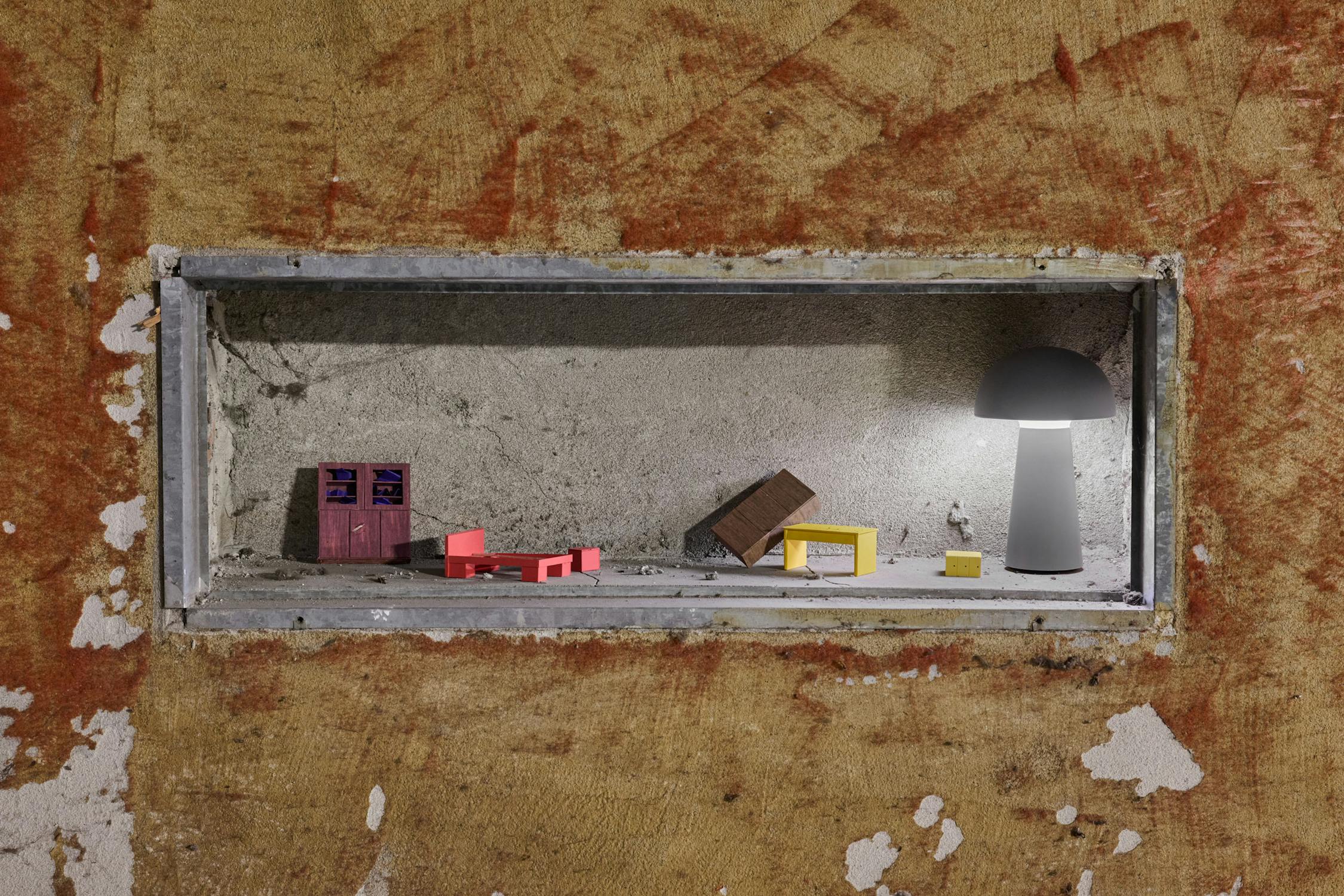 A rectangular niche in a worn, textured wall displays miniature furniture: a pink cupboard, red bed, yellow table, brown bookcase, and a modern grey mushroom-shaped lamp, all arranged on a dusty surface against a rough concrete backdrop.
