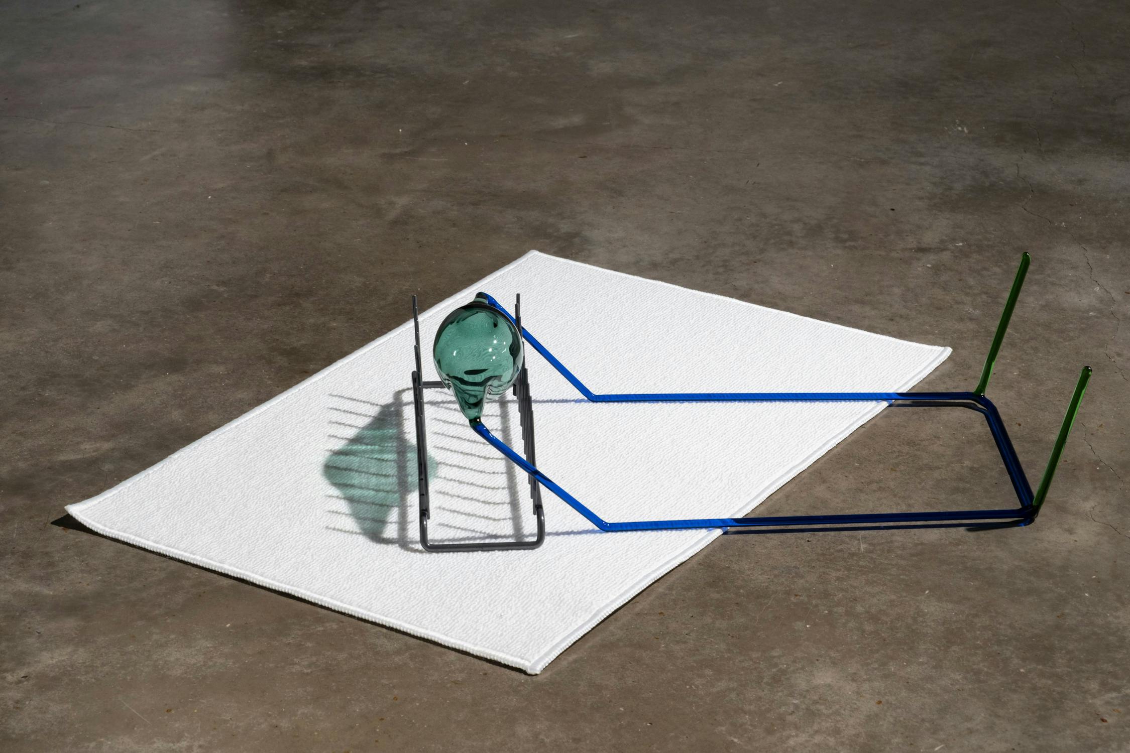 A light glass sculpture head rests upright on a white rectangular mat on a concrete floor. Blue and green glass rods form an angular, abstract frame around the head, casting distinct shadows.