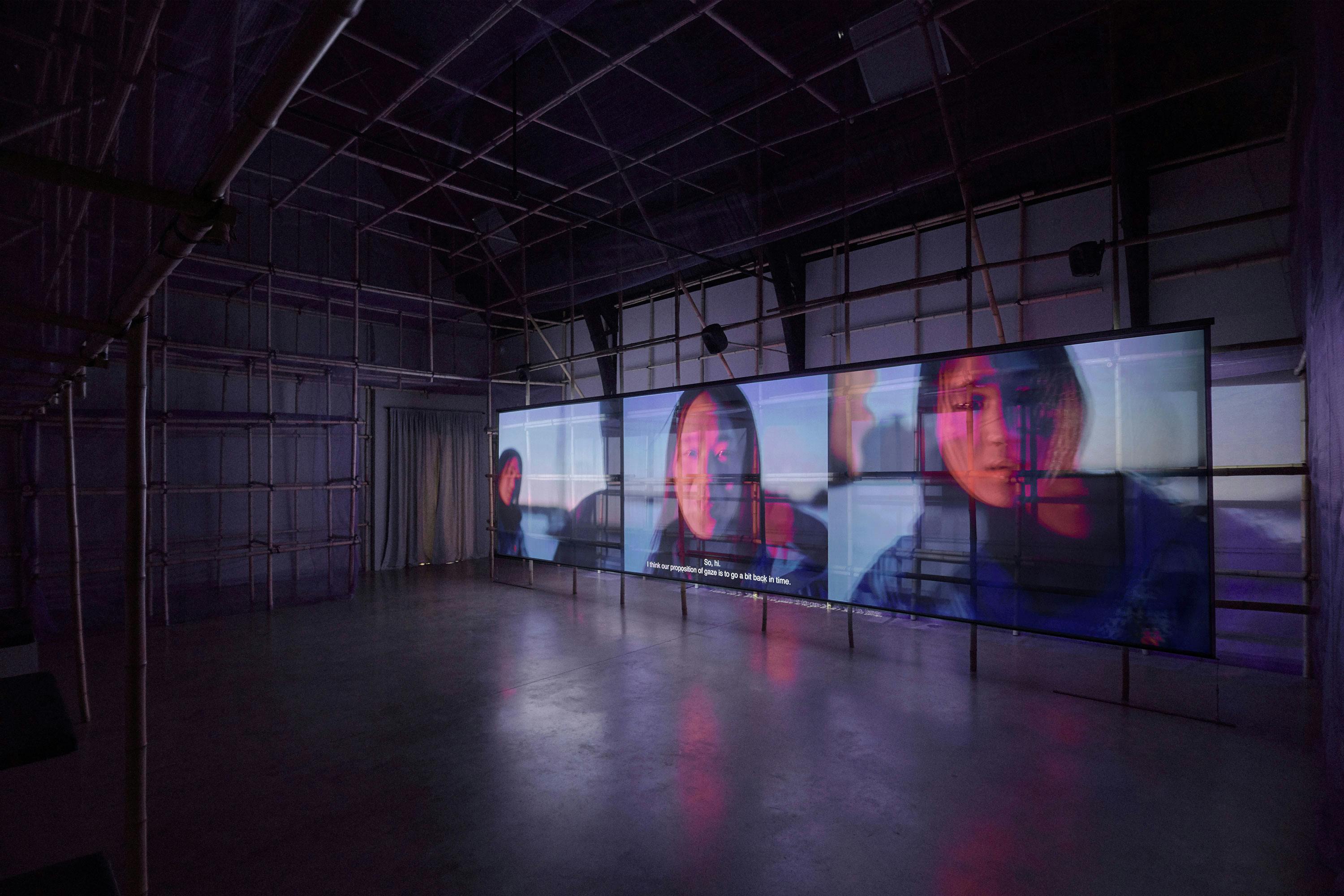 A dimly lit room with three large screens side-by-side showing close-ups of two people, one male and one female, conversing. Subtitles are visible. The room has a grid-like structure on the walls and a reflective, empty concrete floor.