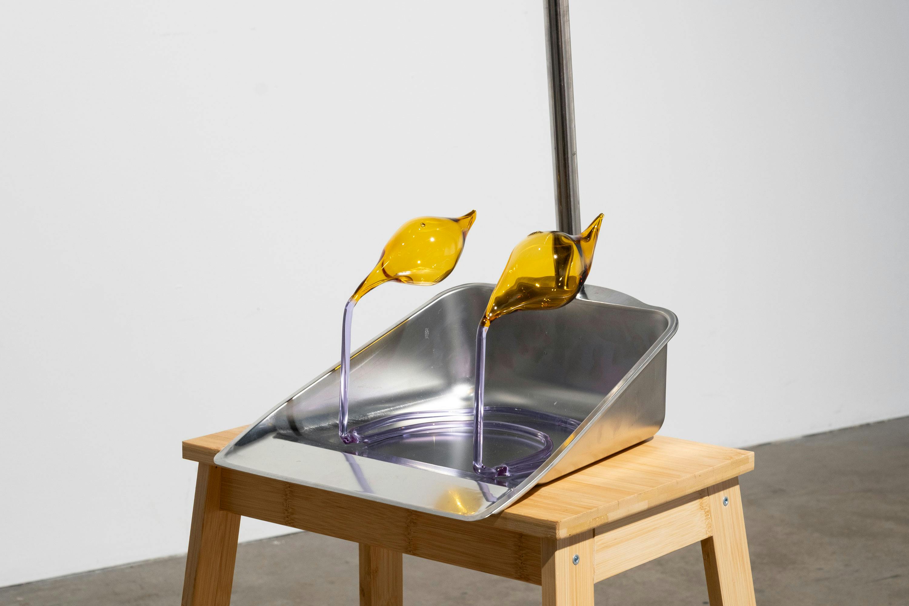 A metal dustpan rests on a wooden stool. Two yellow, bird-shaped glass sculptures with long, transparent necks emerge from the dustpan, creating an abstract, whimsical composition. The background is a plain white wall and concrete floor.