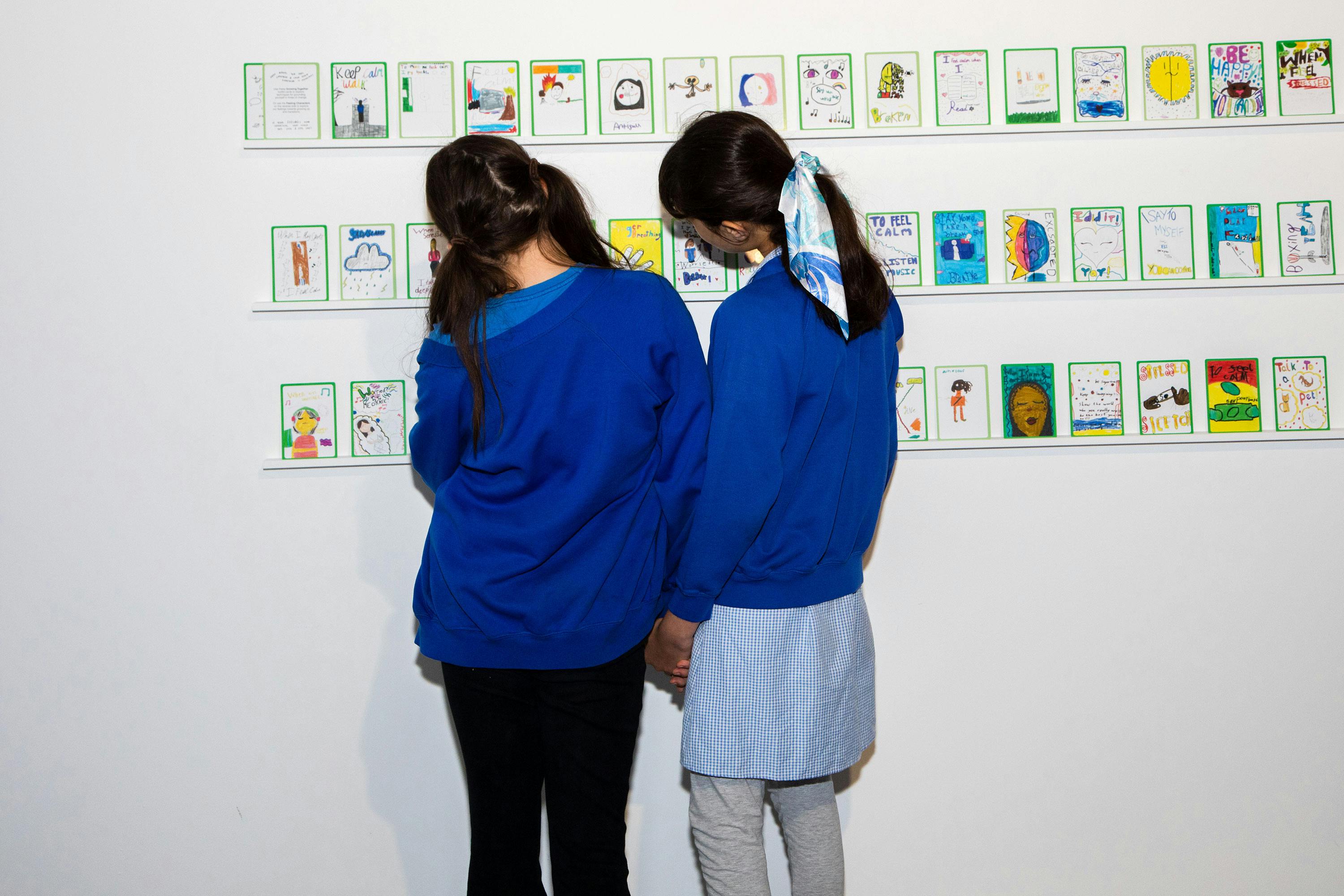 Two children, both wearing blue tops, stand side by side holding hands and looking at a wall display of colourful, childlike drawings arranged in neat rows. The background is a plain white wall, highlighting the vibrant artwork.