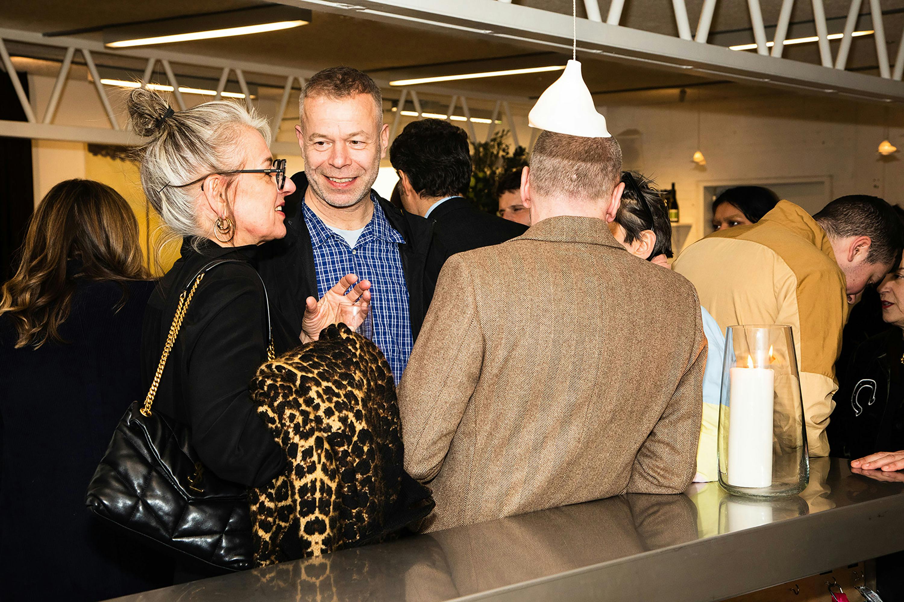 A group of people chat at a busy indoor social gathering. A woman with grey hair and glasses, wearing a leopard-print coat, talks to artist Wolfgang Tillmans. Others stand nearby, including a person in a brown coat and white paper hat, near a large candle in a glass holder.