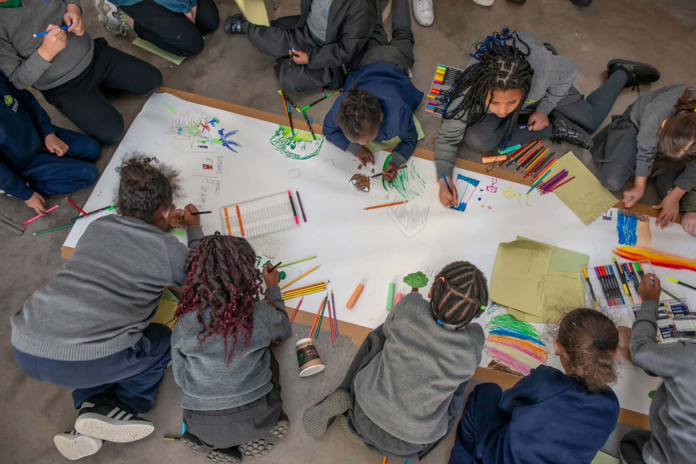 A group of children sit and kneel around a large sheet of paper on the floor, drawing with coloured pencils and markers. The paper is filled with doodles and colourful sketches. Art supplies are scattered around as they work together in a creative activity.