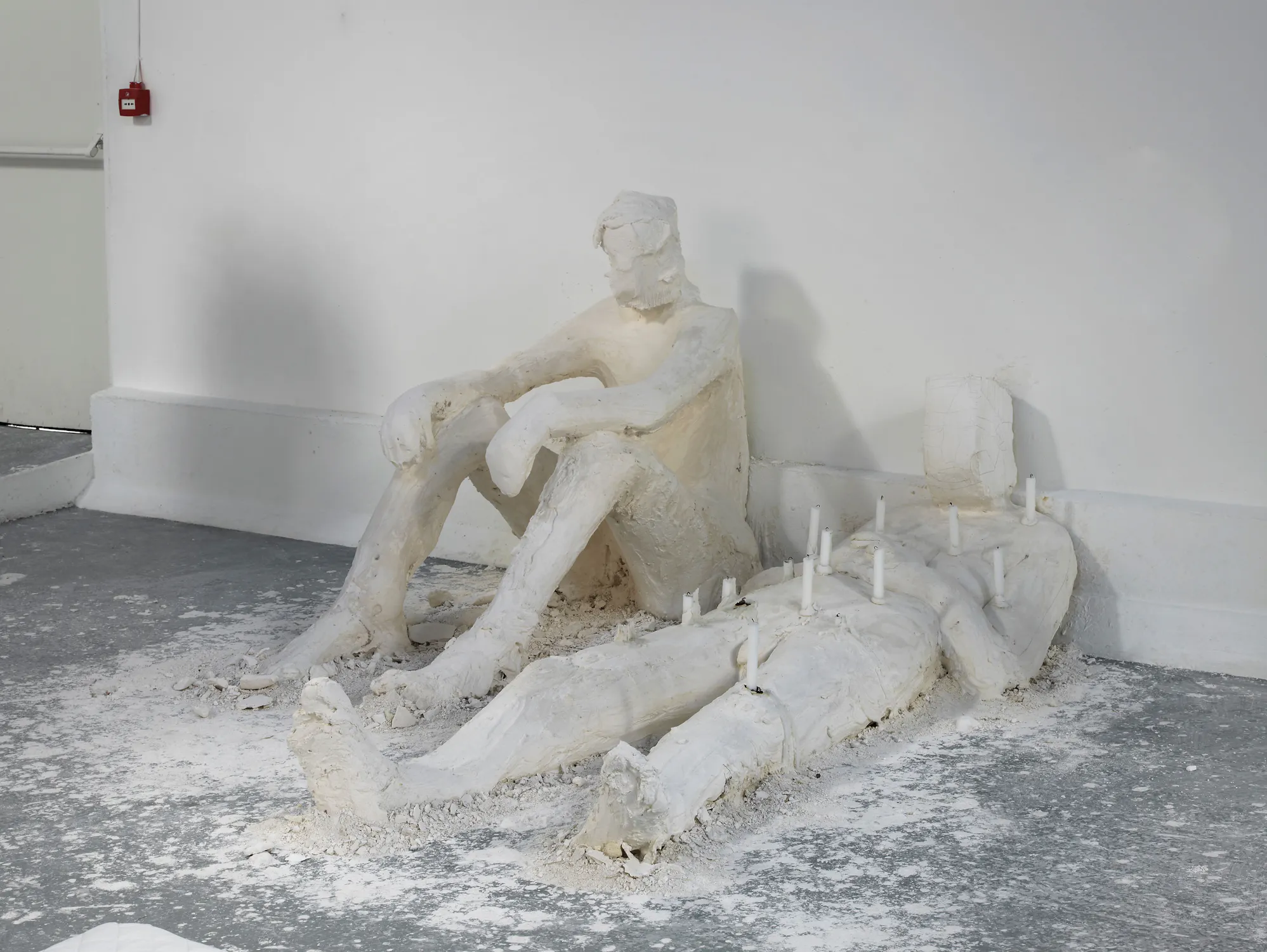 A rough white sculpture by Nicole Eisenman shows a seated figure with elbows on knees, gazing at a reclining figure lying flat on the floor. The prone figure has white candles on its torso, and plaster debris surrounds both forms in a stark indoor space.