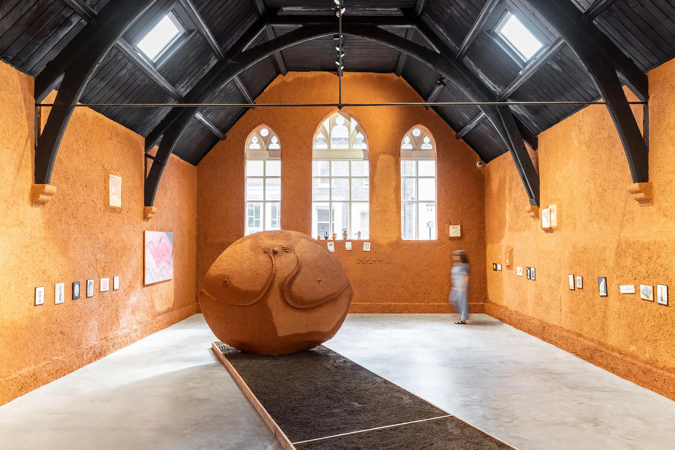 A large, round clay sculpture resembling a reclining human torso sits in the centre of an orange clay-walled gallery with high arched windows. Small artworks hang on the walls and a blurred figure walks near the back, adding a sense of scale.