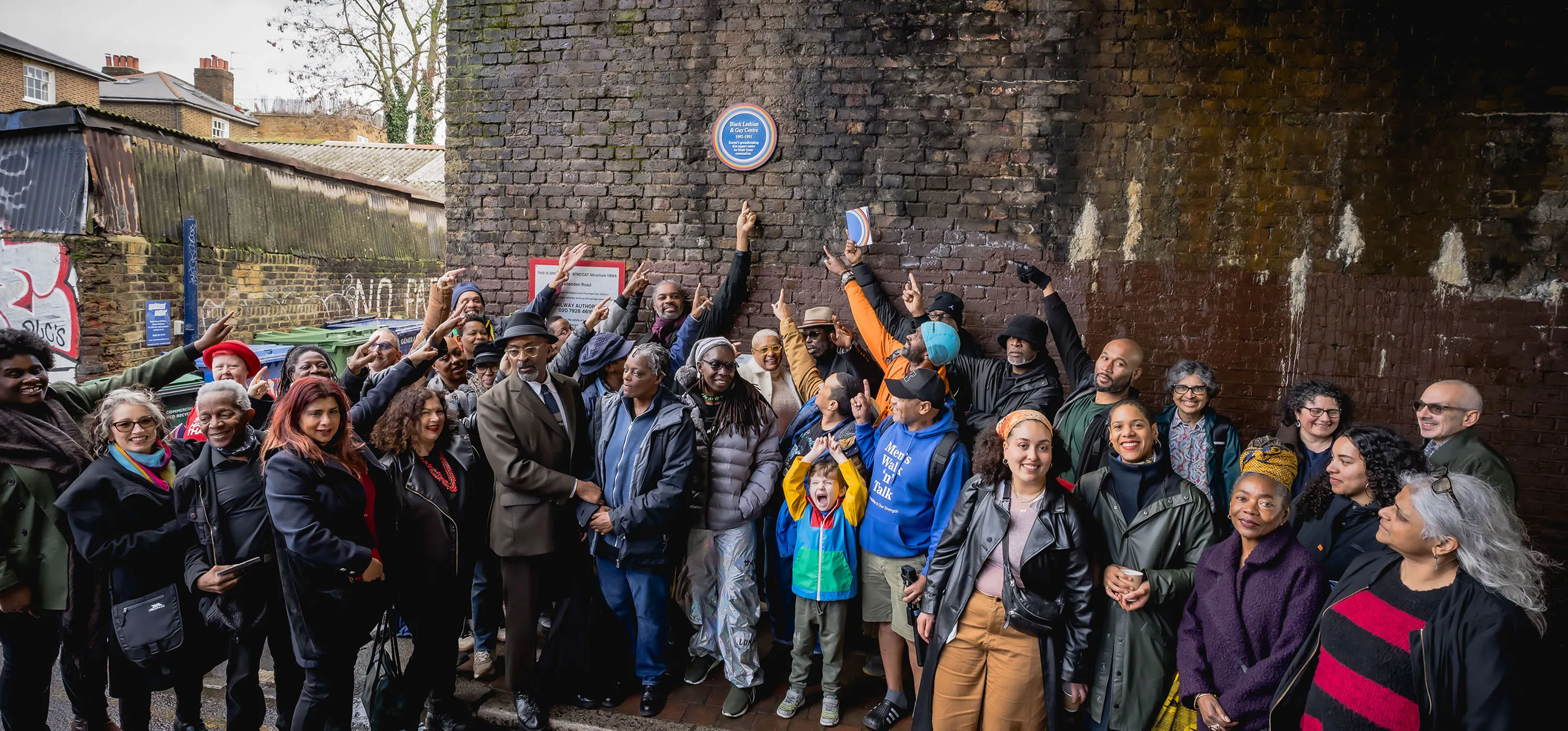 A diverse group of people of various ages and backgrounds gathers under a brick bridge, smiling and pointing up at a blue plaque on the wall. Some hold up mobiles or books, and children stand at the front.