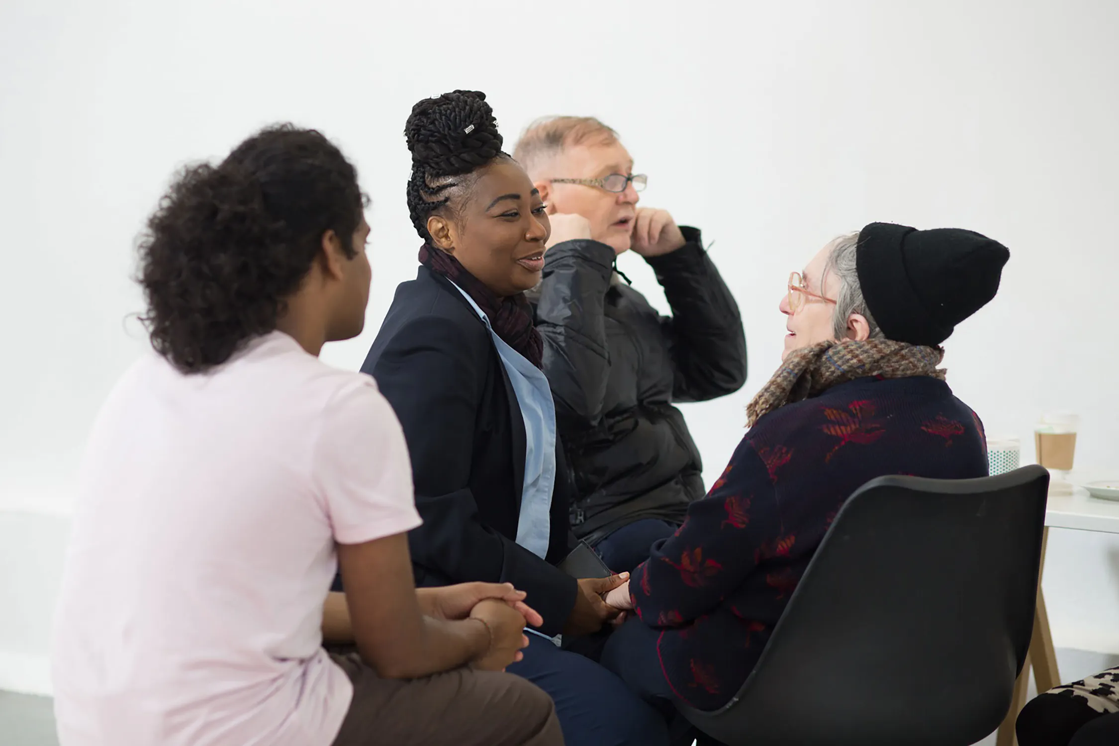 Four people sit in a close circle, engaged in conversation. Two women face each other, one with braided hair and a blazer, the other wearing glasses and a knitted hat. An older man and a young person listen closely. A takeaway coffee cup sits nearby on a white table.