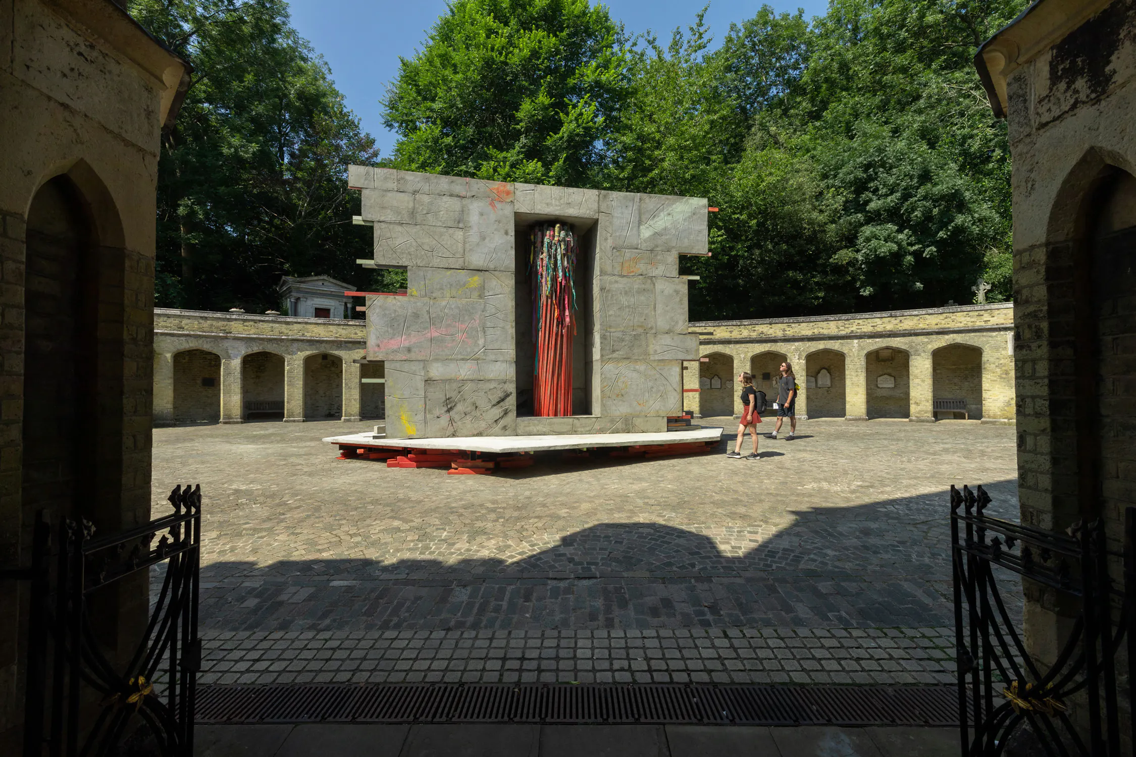 A Phyllida Barlow sculpture depicting a modern stone monument with a vertical red and black art feature stands in a circular courtyard surrounded by stone walls and arches. Two people walk nearby. The scene is shaded by tall green trees in the background.