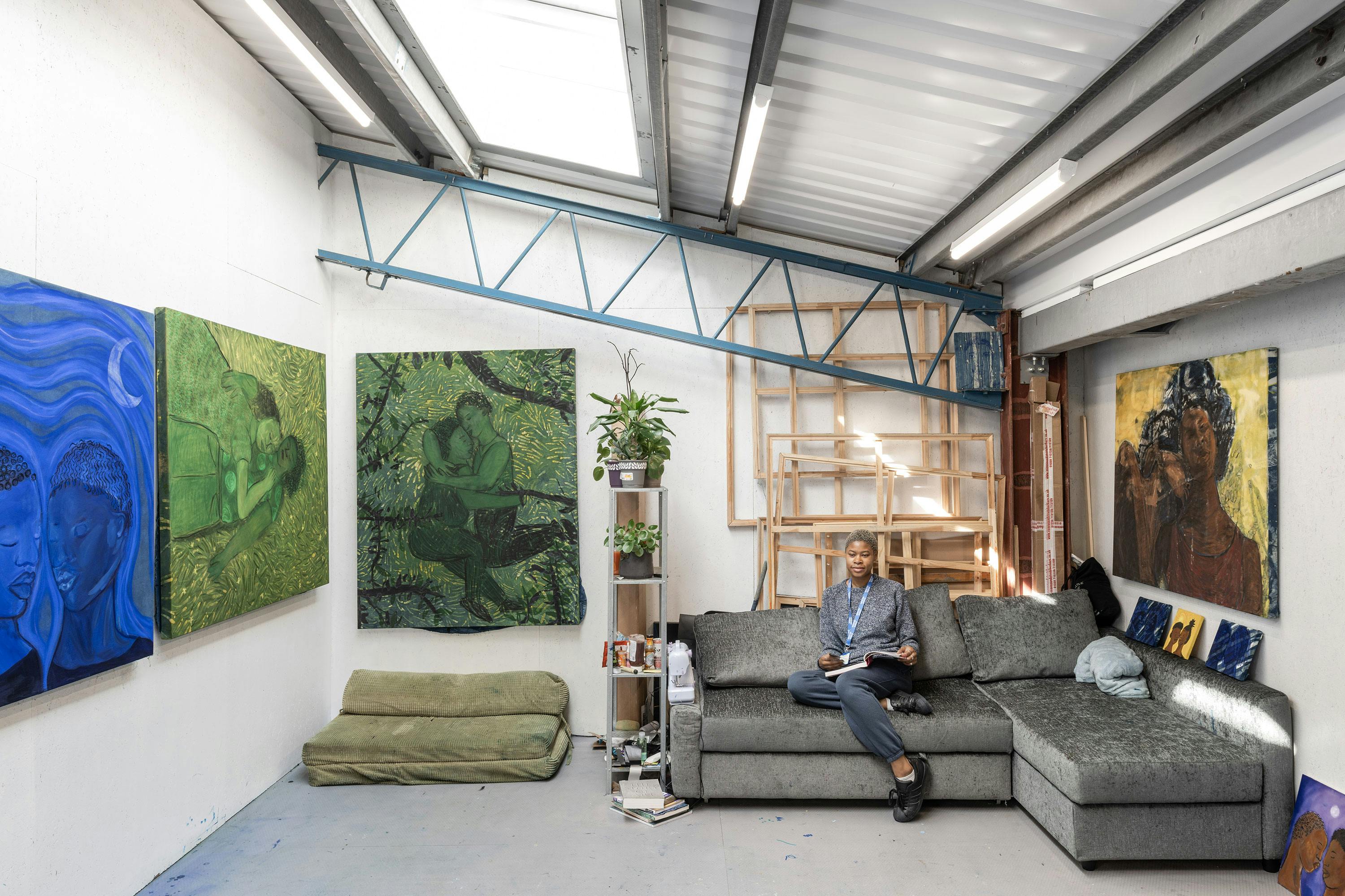 Artist Sola Olulode sits on a grey sectional sofa in a bright art studio. Large colourful paintings, mainly of people, cover the white walls. An easel and stacked canvases are in the corner. A plant and art supplies sit on a small shelf. Lofted ceiling with skylights above.