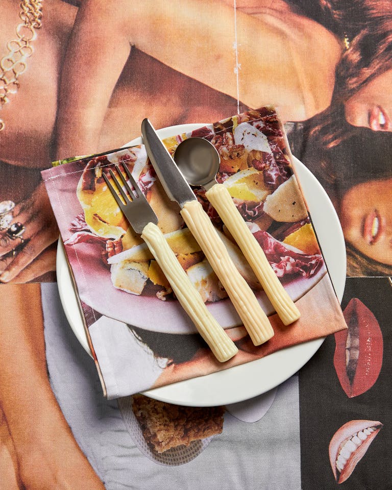 A white plate holds a serviette printed with food, topped with a fork, knife, and spoon with textured cream handles. The plate sits on a collage featuring close-ups of women's faces, lips, and jewellery, creating a bold, layered visual effect.