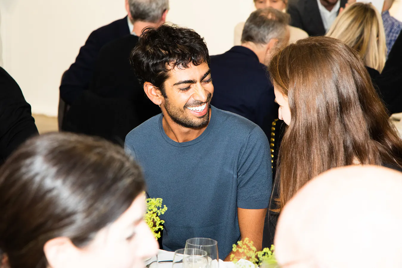 A man with dark hair and a beard, wearing a blue shirt, smiles warmly at a woman with long brown hair at a crowded indoor event. Several people sit around them at tables, conversing and eating.