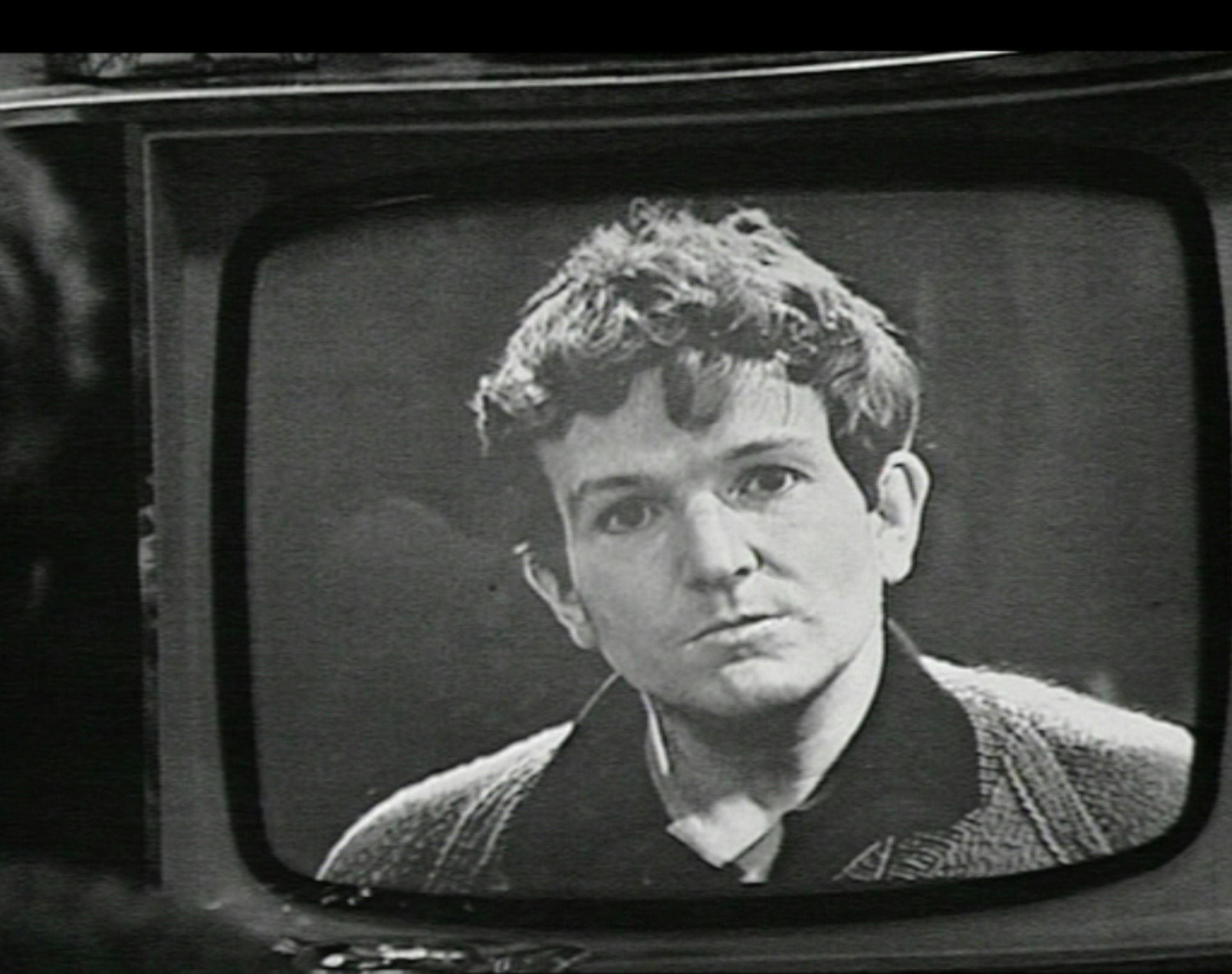Film still of 'Stand Up, Nigel Barton'. A black-and-white image of a man with tousled hair and a serious expression appears on an old television screen. He wears a collared shirt and cardigan. The image is grainy.