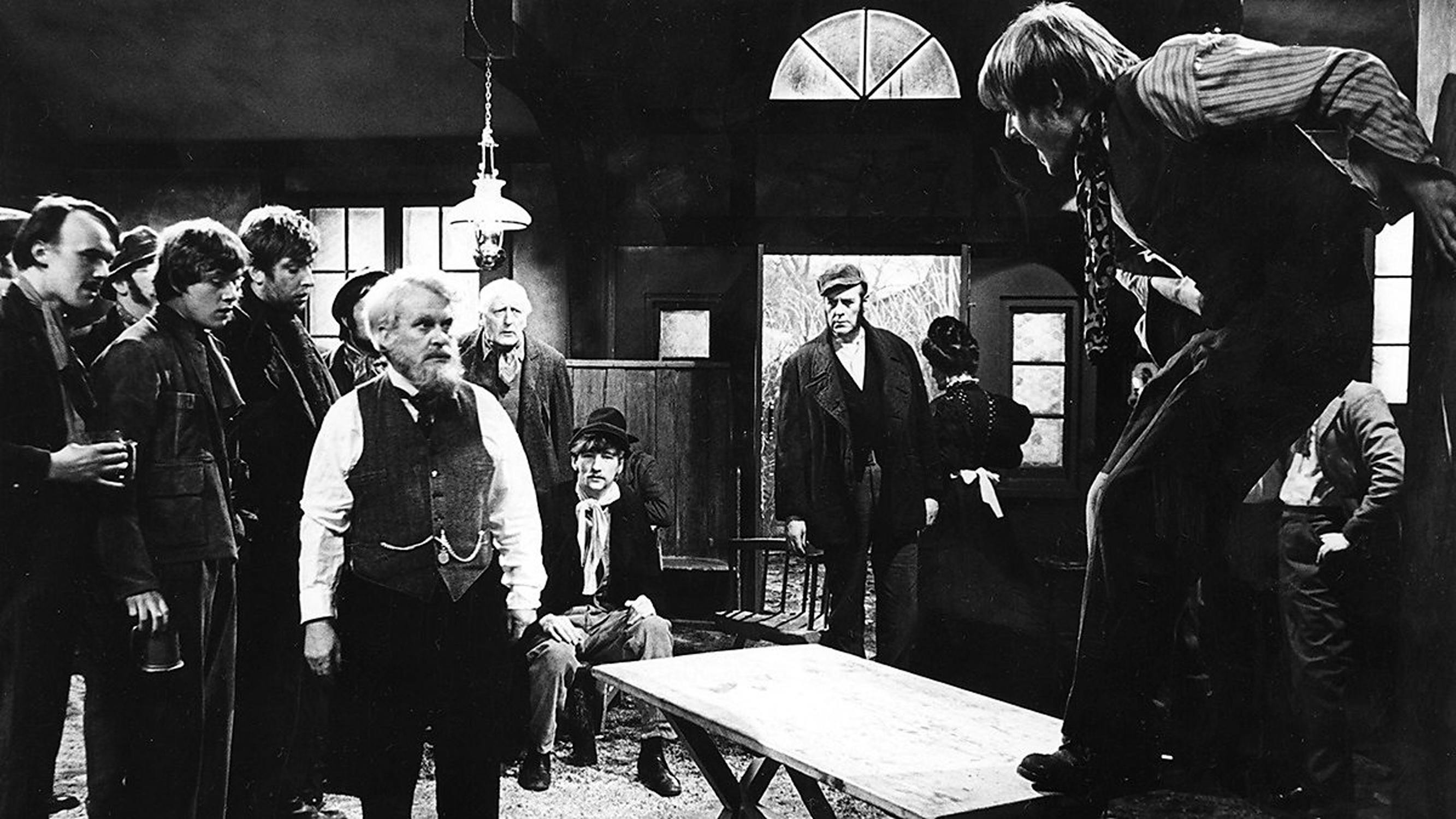 Film still from 'A Beast With Two Backs'. Black-and-white scene shows tense group of men in old-fashioned clothes inside a rustic room. One man stands defiantly on a table, whilst others watch him intently. Lantern hangs from the ceiling; expressions range from concern to curiosity.