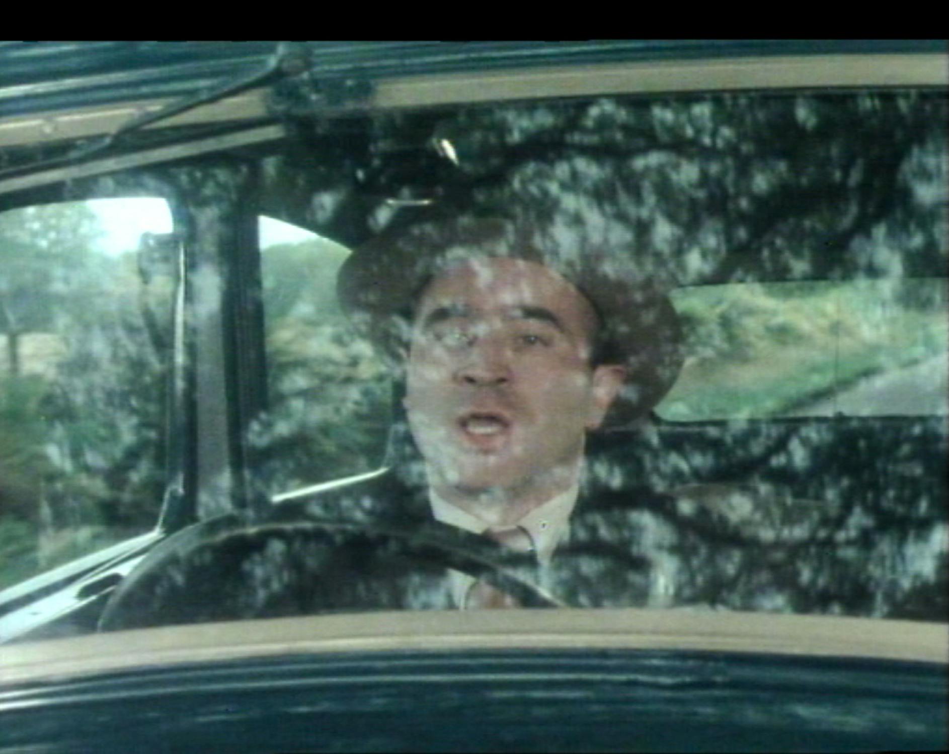Film still from 'Pennis From Heaven'. A man wearing a hat and suit is driving a vintage car, seen through a windscreen splattered with mud or rain. His mouth is open as if talking or singing. Green trees and a rural landscape are visible outside the vehicle.